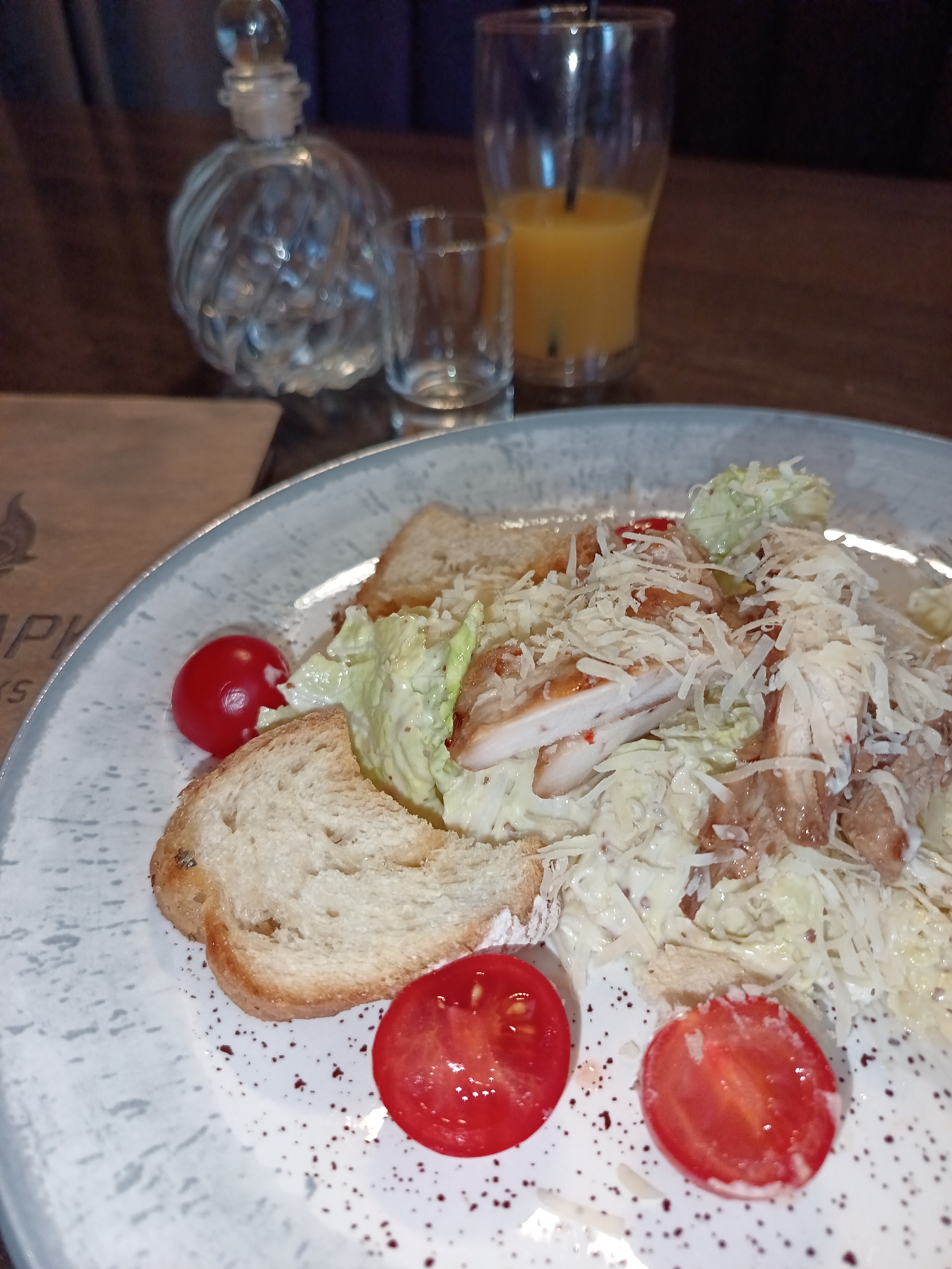 A close-up shot depicts a plate of Caesar salad with grilled chicken, croutons, cherry tomatoes, and shredded Parmesan cheese, set against a dark wooden table. Behind the plate are a decorative glass bottle, an empty shot glass, and a glass of orange juice. The plate features a grey patterned rim and is speckled with dark specks. The photograph appears to be taken in a restaurant setting, possibly in the city of Osa, Russia. There are no people visible in the shot. The focus is on the arrangement of the Caesar salad and the accompanying beverages.