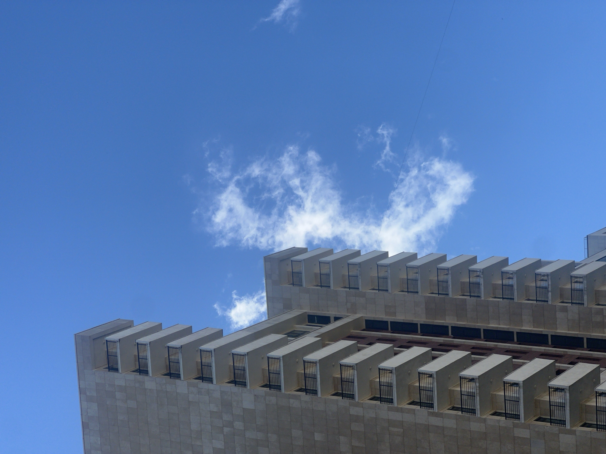 The image shows a modern apartment building with multiple balconies against a bright blue sky with scattered white clouds. The building has a light-colored, textured facade and dark railings on the balconies. The architectural style suggests it is a relatively new construction. There are no people visible in the image. The scene is a clear, sunny day. The location is not identifiable as Moscow, Russia from the visual information provided.
