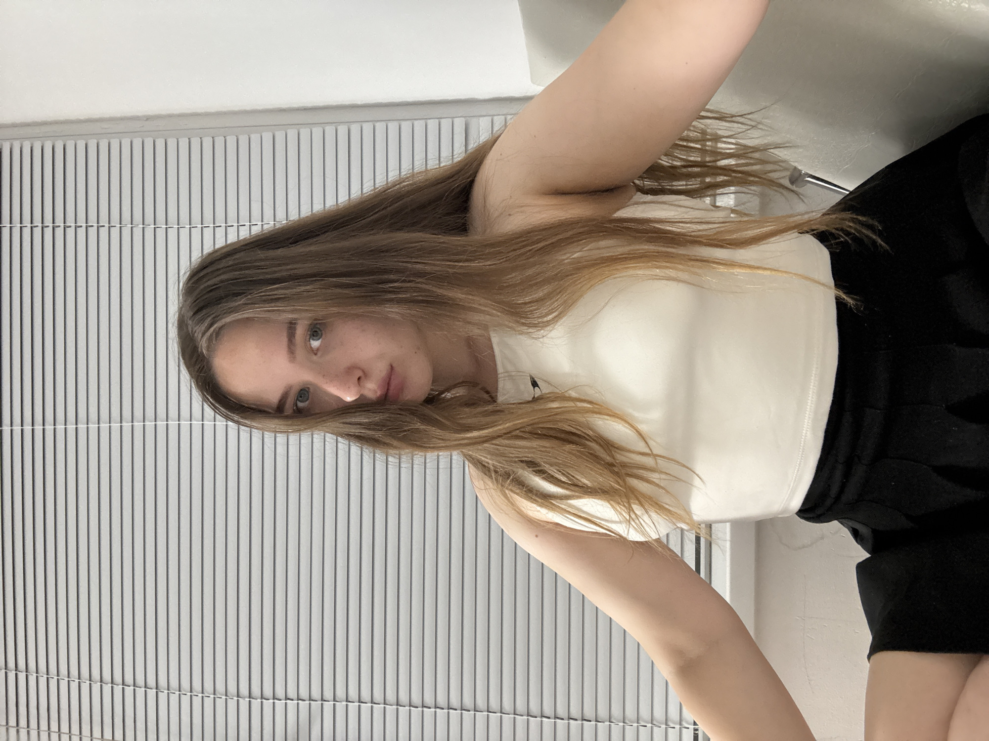The media file shows a close-up, sideways shot of a young woman with long, light brown hair and fair skin, looking towards the camera. She is wearing a white tank top and black shorts. Her facial expression is neutral. Her hair is slightly messy and falls around her face and shoulders. The background consists of vertical blinds, suggesting an indoor setting. The lighting appears to be artificial, and there are no clear indicators of the time of day or weather conditions. The image is a selfie, with the camera angled from above and to the side. No text is visible in the image. The setting suggests a casual, indoor environment, possibly a home or studio.