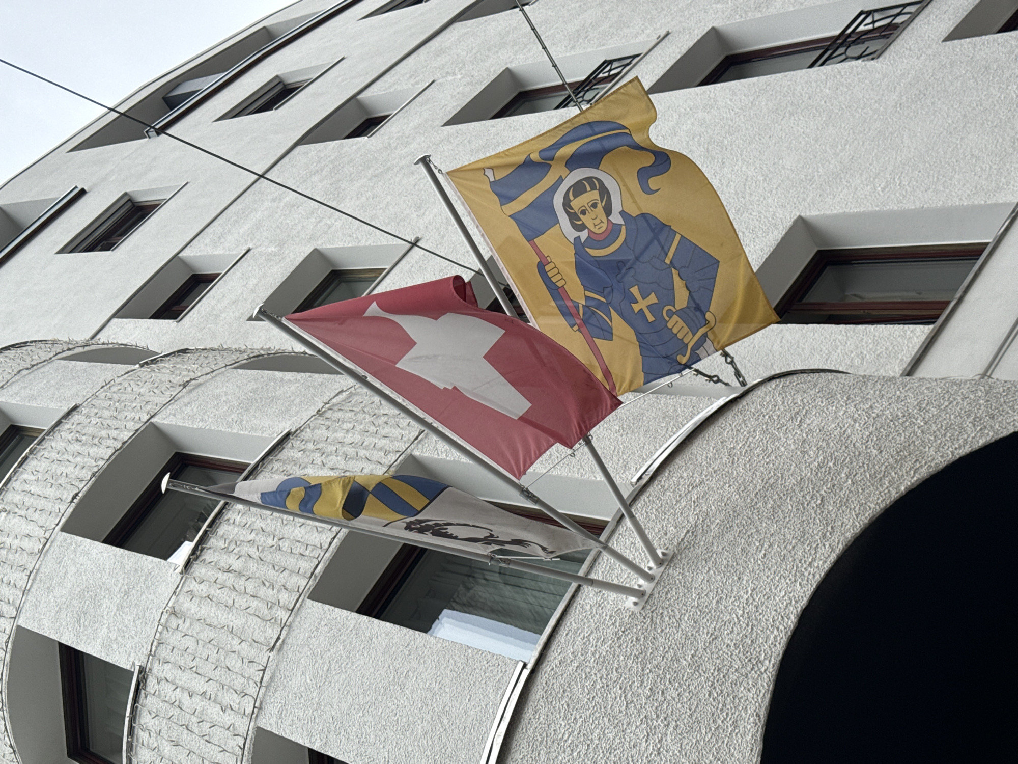 This is a low-angle, outdoor, medium shot of three flags flying from the side of a white, modern building. The flags are mounted on a white bracket attached to the building. The building has several windows with dark frames. The flag on the left is white with a black, circular emblem. The middle flag is the Swiss flag, which is red with a white cross. The flag on the right is yellow with a blue and black depiction of a knight in armor holding a lance and a shield with a cross. The background is an overcast sky. The flags appear to be in the city of St. Moritz, Switzerland, a well-known alpine resort town in the Engadin valley.