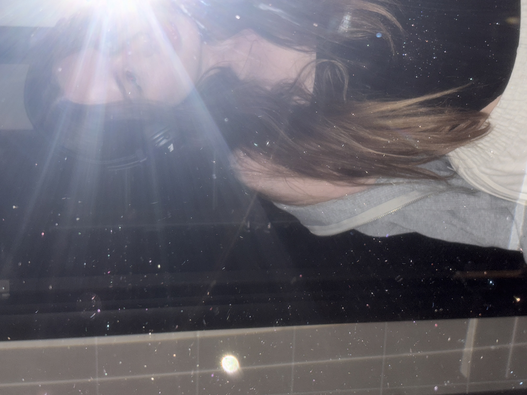 In a dimly lit indoor environment, a young woman is partially visible, facing a reflective surface. She is wearing headphones and a grey jacket over a black top, with her long, light brown hair cascading down. A bright light source, possibly a flash, is positioned to her right, creating lens flare and obscuring parts of her face and the background. The reflective surface appears to be a mirror, exhibiting numerous small, scattered dust particles or glitter, some of which are reflecting the light with subtle iridescent hues. The background is dark and indistinct. The foreground features a tiled wall on the left side of the image, with a circular light reflection on it. This scene is not identifiable as being located in Chaikovsky, Russia.