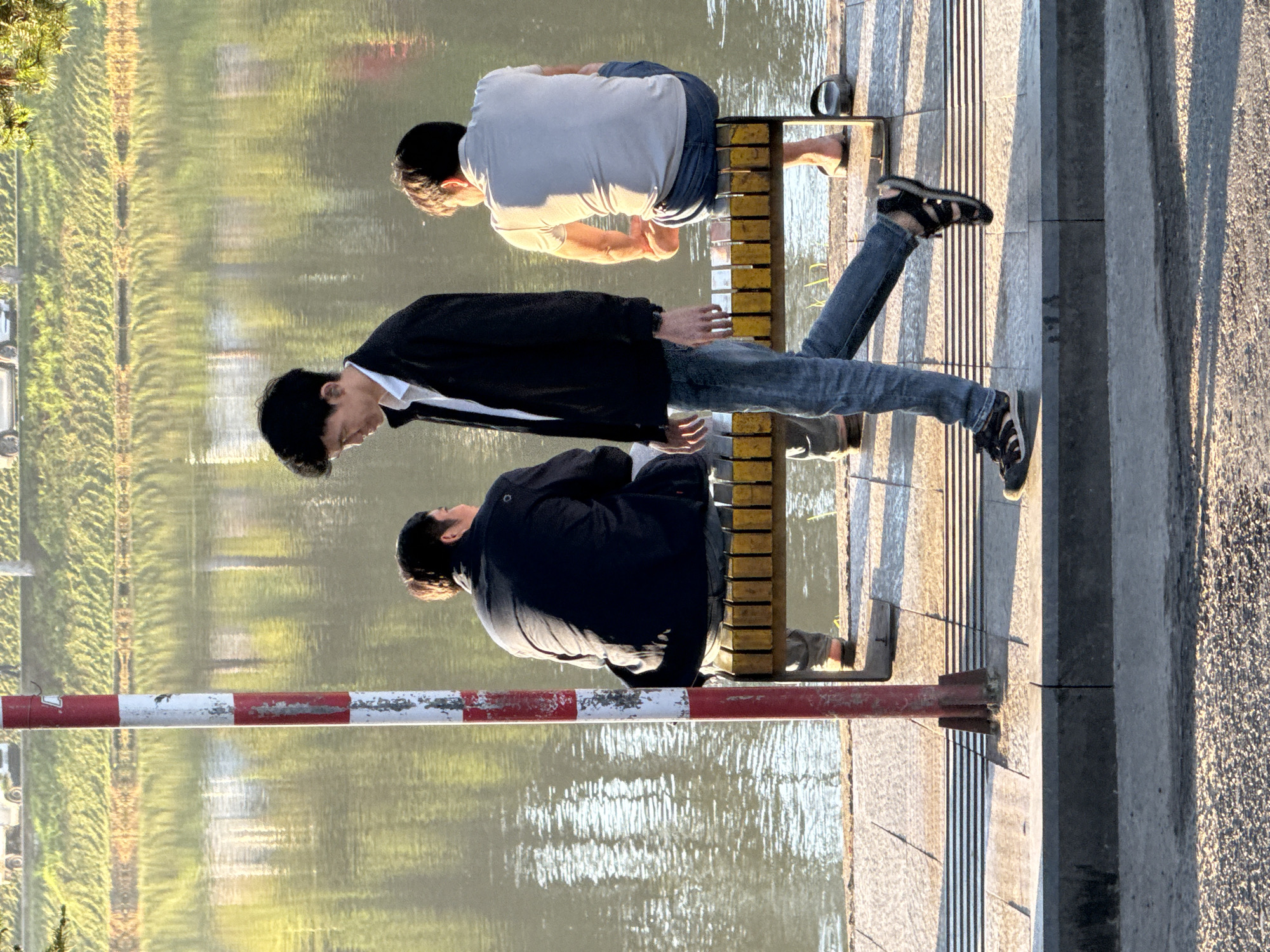 This image captures a scene along Xuan Huong Lake in Da Lat, Vietnam, during daylight hours. Three individuals are present. In the foreground, a young man walks from left to right, wearing a dark jacket over a white shirt and blue jeans, with dark sandals on his feet. To his left, a person is seated on a bench, facing away from the camera, wearing a dark jacket and dark pants. To the right of the walking man, another person is seated on the same bench, also facing away, dressed in a light-colored t-shirt and blue jeans, with their feet bare. The bench is wooden with yellow slats. A red and white striped pole stands to the left of the frame, partially obscuring the seated figures. Behind the bench and pole is the tranquil, reflective surface of Xuan Huong Lake, with greenery and buildings visible in the distance on the opposite shore. The ground in the foreground is paved with textured tiles and asphalt. The overall lighting suggests it is late afternoon, with the sun casting long shadows.