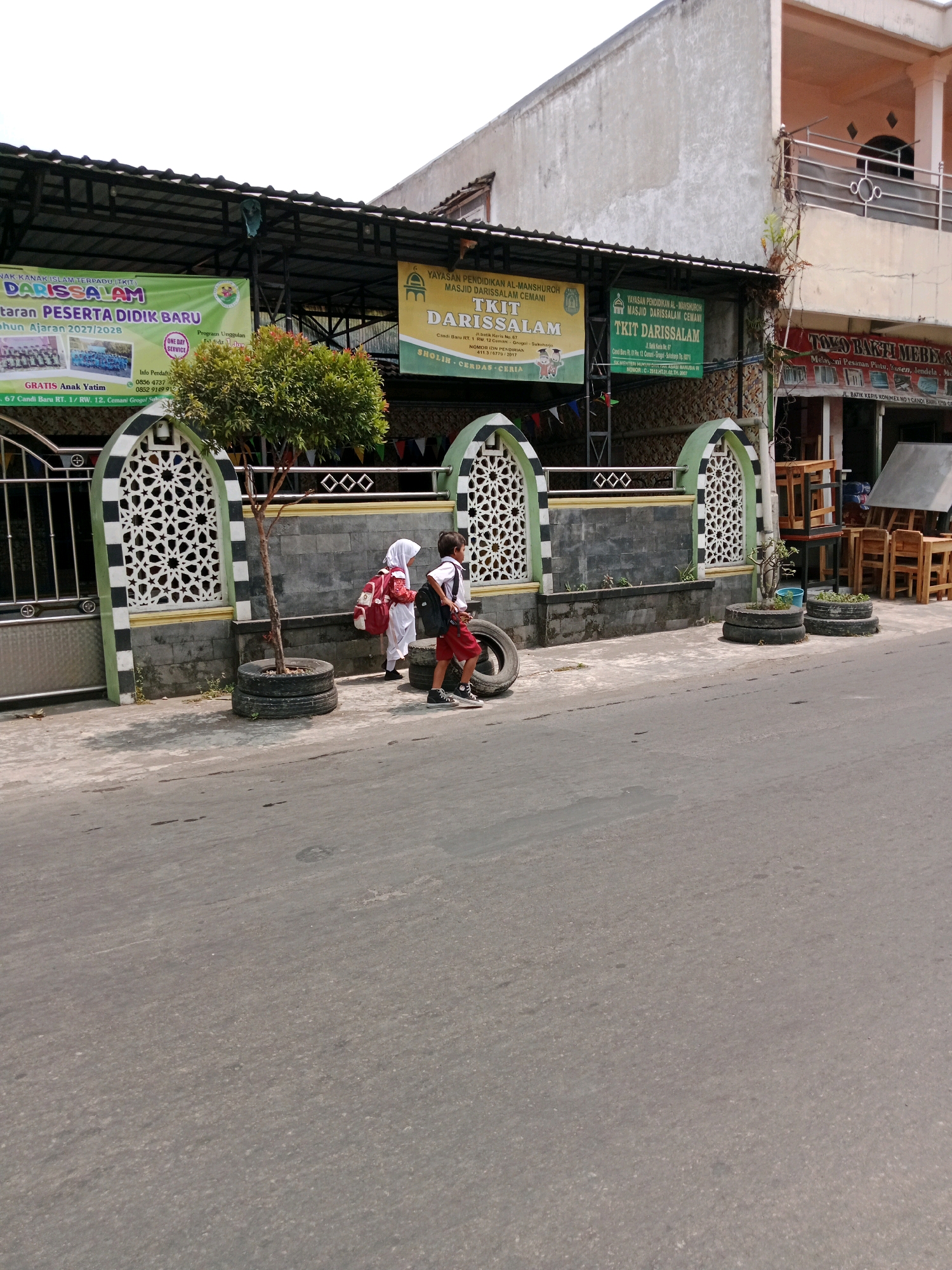 The image depicts the street view of a school building in Cemani, Indonesia. Two children, a boy and a girl wearing school uniforms, are walking on the sidewalk in front of the school. The boy is wearing red shorts and a white shirt, carrying a black backpack, and holding a tire. The girl is wearing a white hijab and a white dress, also carrying a backpack. The school building has a green and white facade with decorative patterns on the wall. Banners with school information are visible above the entrance. The street is paved with asphalt, and there are some trees and potted plants along the sidewalk. The scene appears to be during daytime, with bright sunlight casting shadows. There are also some wooden furniture stacks on the right side of the image, possibly from a nearby shop.