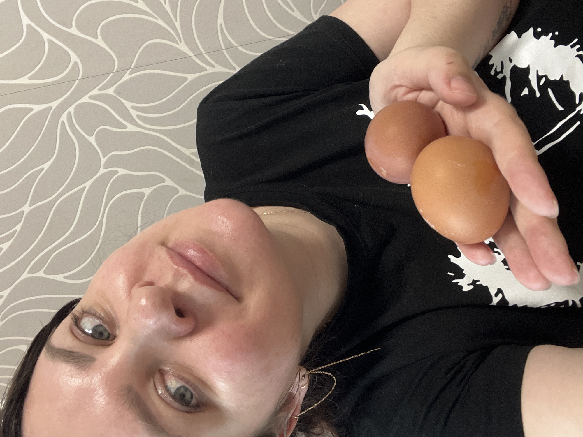 A medium close-up image shows a person, likely a woman, holding two brown eggs in their right hand and looking towards the camera. The person's head is on the left side of the frame, tilted slightly upward, with their body extending diagonally to the right. They have blue-gray eyes and a soft, neutral expression with a hint of a smile. Dark hair frames their face, and a delicate gold chain earring is visible on their right ear. A thin gold chain necklace is also visible around their neck.
The individual is wearing a black t-shirt featuring a prominent white abstract or splash-like graphic design on the chest. The background is a wall with light gray wallpaper patterned with flowing, organic lines in a lighter shade, possibly off-white or very light gray. The lighting suggests an indoor setting. No specific time of day or weather conditions can be determined. The image appears to be a casual, self-portrait style photograph, possibly taken from a low angle. The location context for the scene is Perm, Russia. No discernible text is visible in the image.