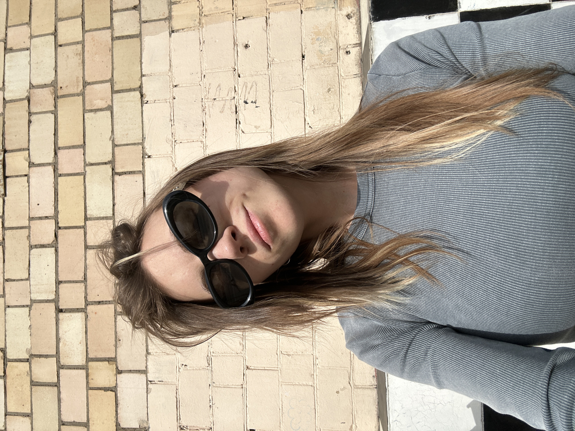 A young woman with long, blonde hair is photographed from a high angle. She is wearing dark sunglasses and a grey, ribbed, long-sleeved shirt. Her lips are slightly parted, and she appears to be looking up and to her left. The background is a light-colored tiled wall. To the right of the woman, there is a black and white checkered surface. The lighting suggests it is daytime, and the woman's hair is illuminated by sunlight, indicating fair weather. The presence of tiles and the checkered pattern suggest an urban or possibly indoor setting, consistent with a location in Moscow. There is no visible text in the image.