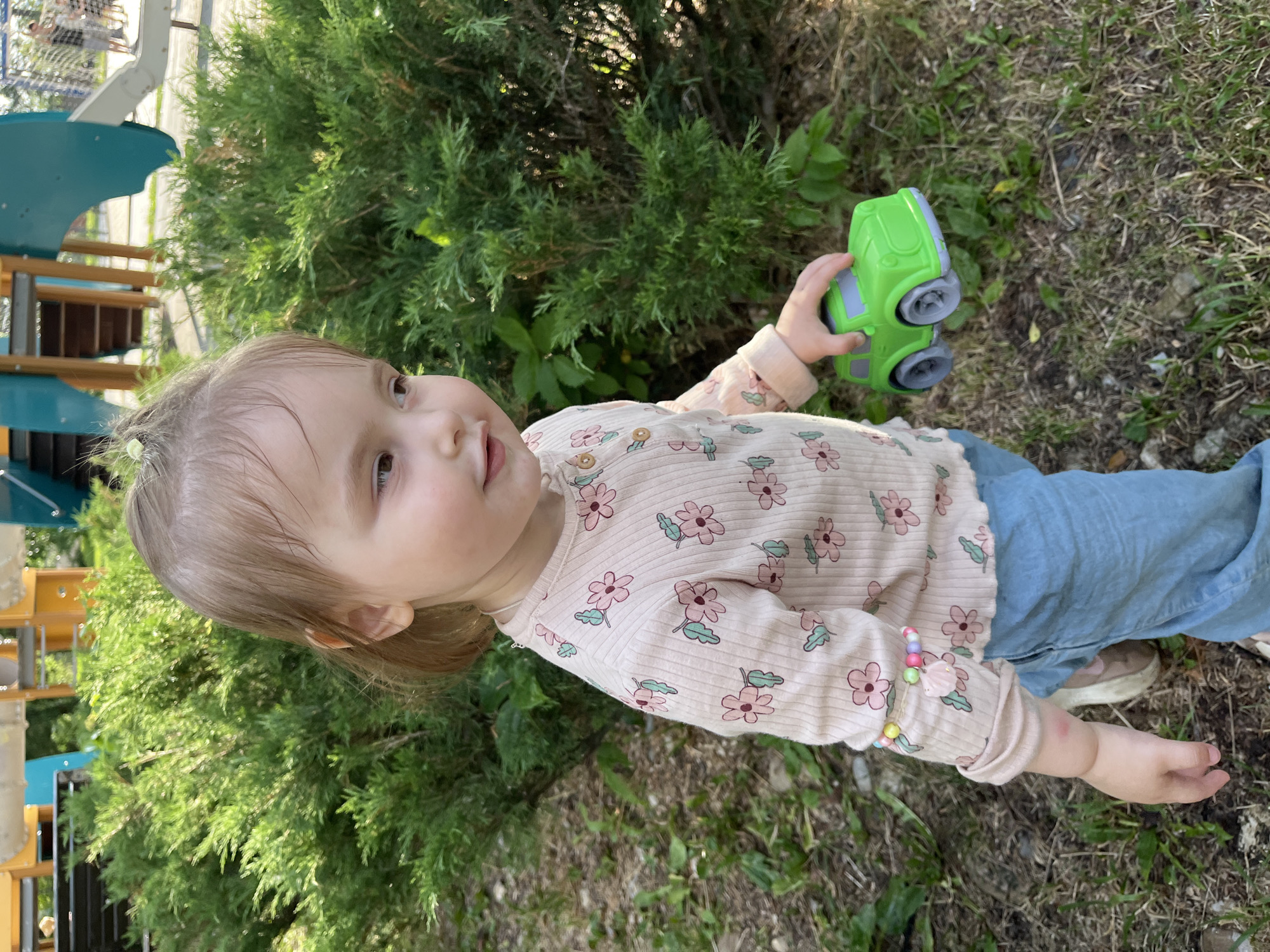 A young Caucasian girl stands outdoors, holding a green toy car. She is wearing a light pink long-sleeved shirt with a floral pattern and light blue jeans. She has light brown hair and is looking to her right. The background consists of green bushes and a playground structure, suggesting a park or recreational area in Novokuybyshevsk, Russia. The ground is covered with grass and some fallen leaves.