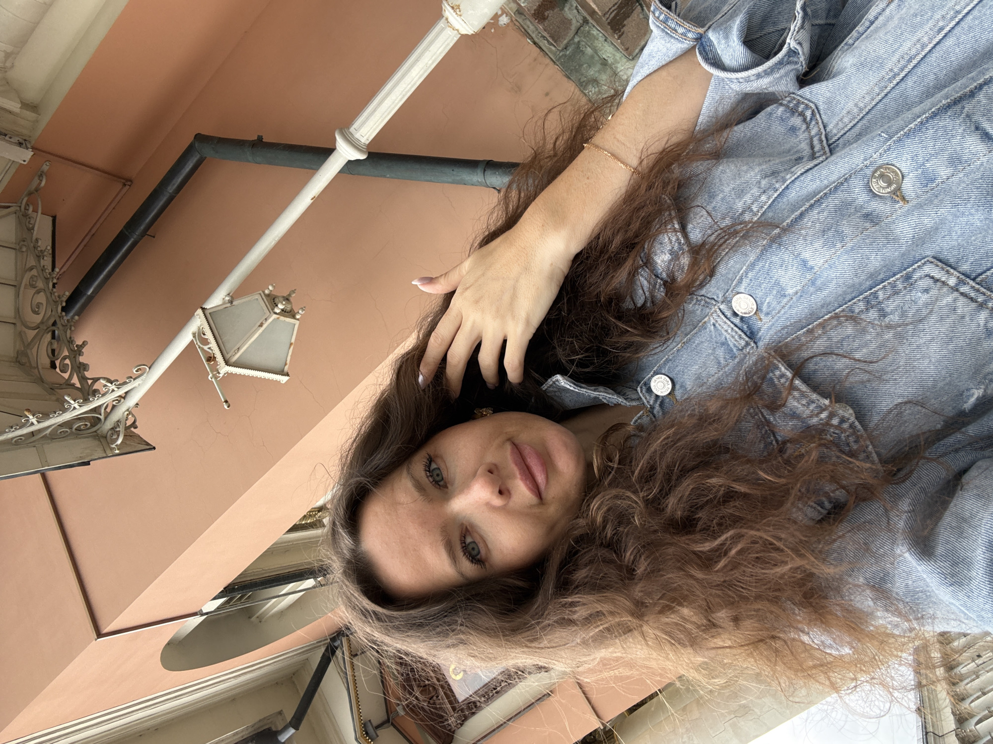 A young woman with long, wavy brown hair is taking a selfie outdoors. She is wearing a light blue denim jacket. Her right hand is raised to her head, touching her hair. The background shows a pale pink building with arched windows, an ornate white lamppost, and a dark downspout. The lighting suggests it is daytime. The location is unidentified.