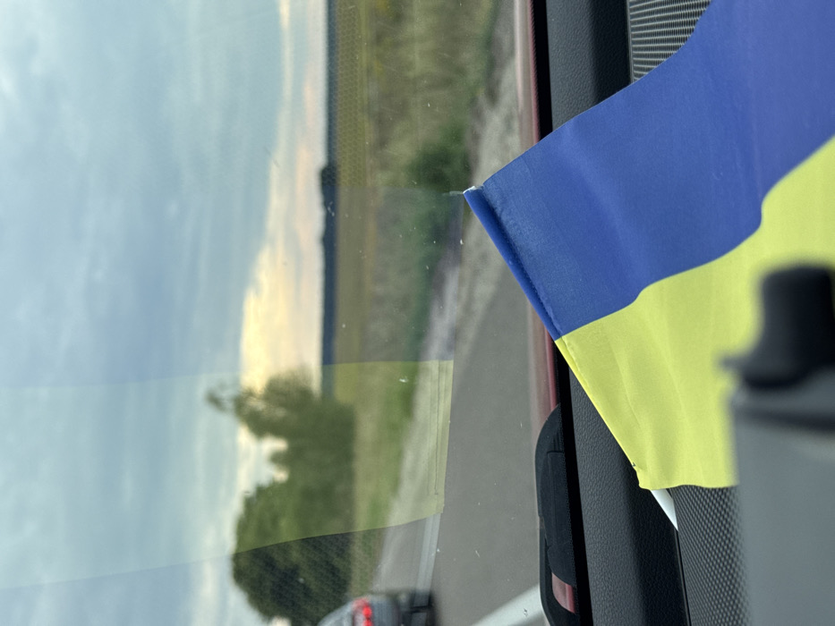 The image depicts a view from the interior of a vehicle traveling on a road in Bezuhlivka, Ukraine. In the immediate foreground, on the right side of the vehicle's dark-colored dashboard, a Ukrainian flag is partially visible, displaying its blue upper band and yellow lower band. The flag appears slightly crumpled. Through the windshield, a two-lane asphalt road with a solid white line on the left is visible. Another dark-colored vehicle, likely a sedan or hatchback, is seen ahead on the road, partially showing its rear and red taillights. The roadside on the left features a dense line of green trees and sparser light-green foliage. To the right of the road, expansive agricultural fields, appearing yellow-green and extending towards the horizon, dominate the landscape. The sky is overcast, with shades of grey clouds, and a soft, diffused yellowish-orange light is present near the horizon line. No individuals are visible.