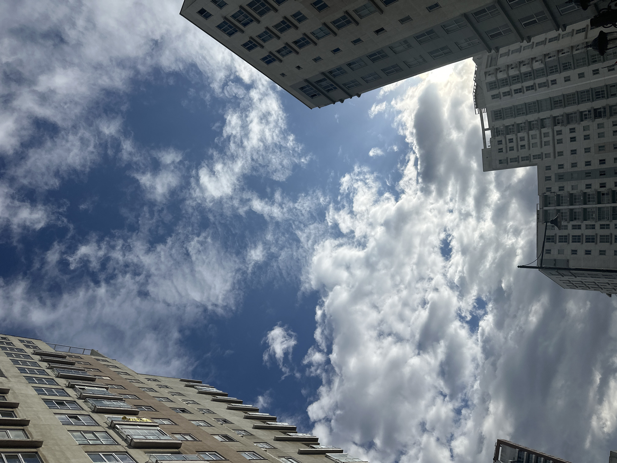 The image is a low-angle shot of buildings and the sky in Esenyurt, Turkey. The sky is mostly blue with scattered white clouds. The buildings, which appear to be residential, are of different heights and architectural styles. One building is gray, another is white. Sunlight is visible breaking through the clouds. There are no people visible in the image.