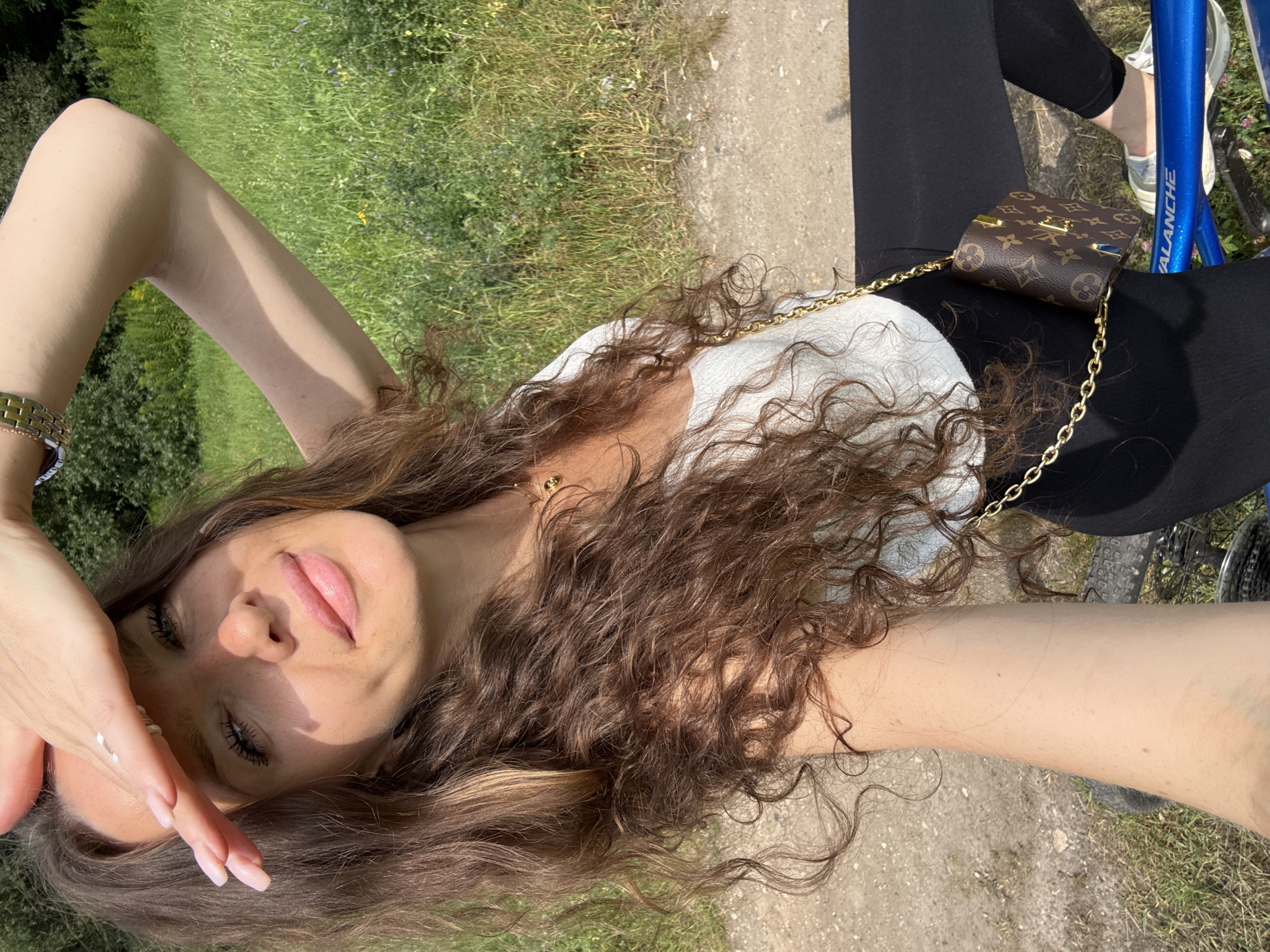 A woman with long, curly brown hair is posing for a selfie outdoors on a sunny day, possibly in Moscow, Russia, given the presence of a person wearing a wristwatch. She is wearing a white, ruffled top, black leggings, and light blue sneakers. She is holding her right hand up to shield her eyes from the sun. A small brown designer purse with a gold chain is hanging from her shoulder. She is seated on a blue bicycle. The background consists of green grass and trees.