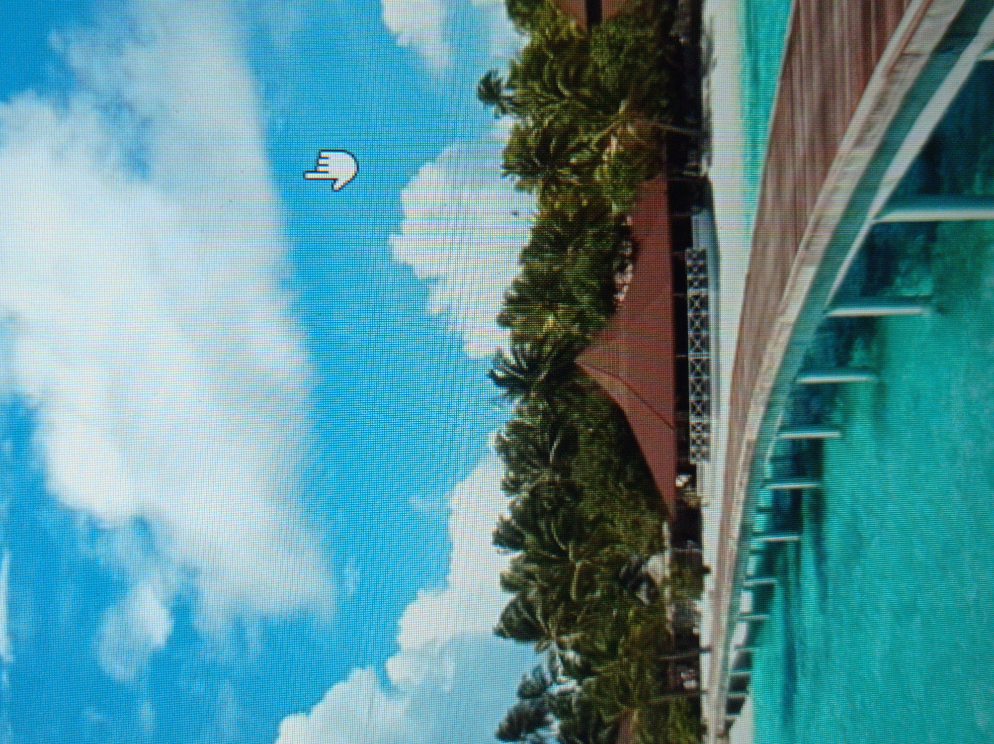 The image depicts a tropical island scene with a pier extending over turquoise water. In the background, there is a beach lined with palm trees and a building with a reddish-brown roof. The sky is blue with fluffy white clouds. A small white hand cursor icon is visible in the upper right corner of the image, indicating a digital interface. This image is not related to the city of Mukachevo, Ukraine.