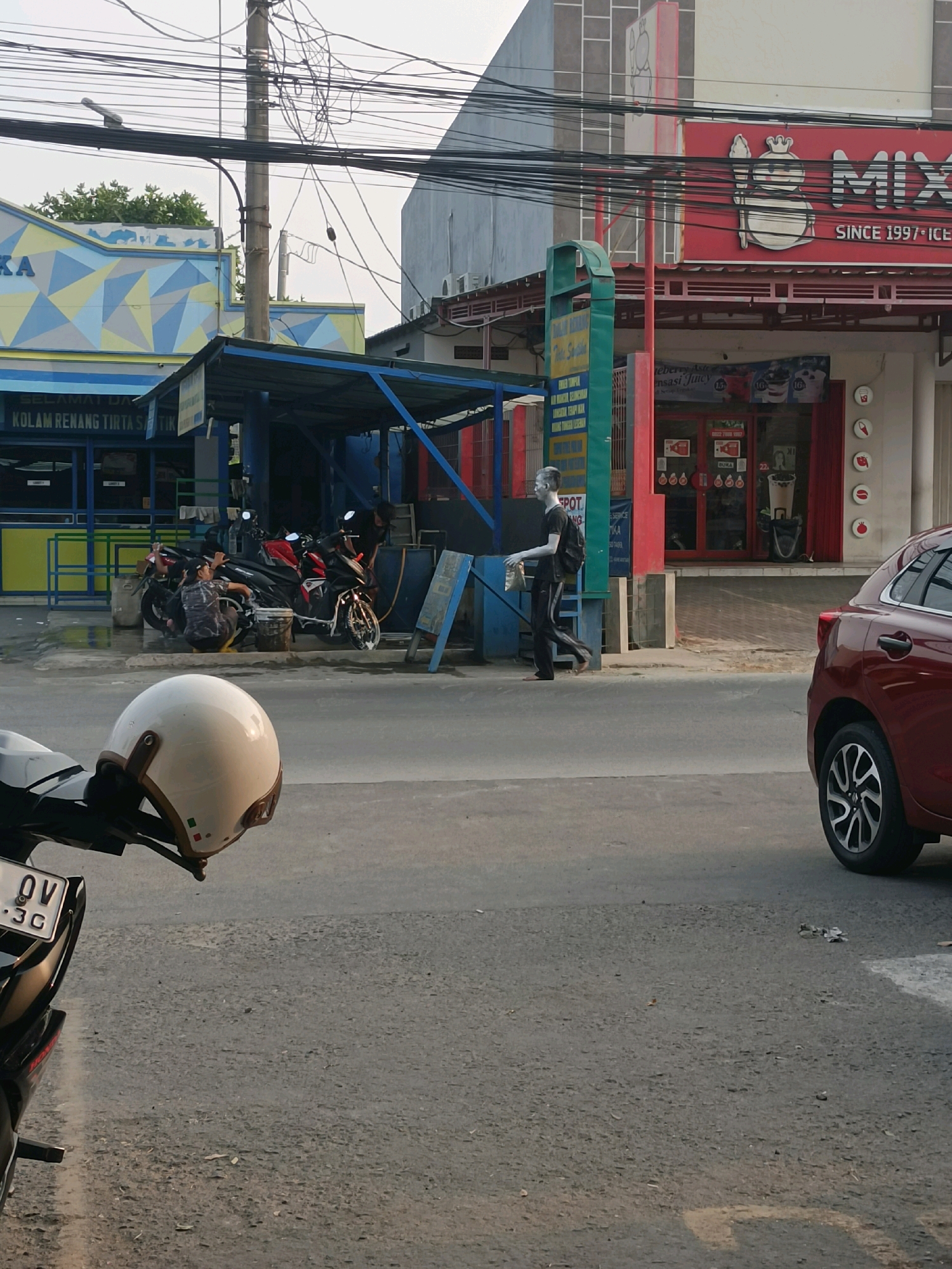 The image depicts a street scene in Karawang, Indonesia, likely in a commercial area. In the foreground, a white helmet rests on the handlebars of a black motorcycle. The motorcycle's license plate displays "OV .3G".
Further down the street, several motorcycles are being washed under a blue-roofed shelter. Two individuals are engaged in washing the motorcycles. To the right of the shelter, a man with a light-colored, possibly painted, face and a black backpack walks past. He is wearing dark pants and appears to be carrying a plastic bag.
Behind the shelter and the motorcycle washing area, a red building with a prominent "MIX" sign and "SINCE 1997 ICE" text is visible. Adjacent to this is another building with a blue and yellow geometric facade, labeled "KA". Power lines crisscross overhead, supported by a utility pole. A red car is parked on the right side of the street. The environment appears to be a roadside with paved asphalt.