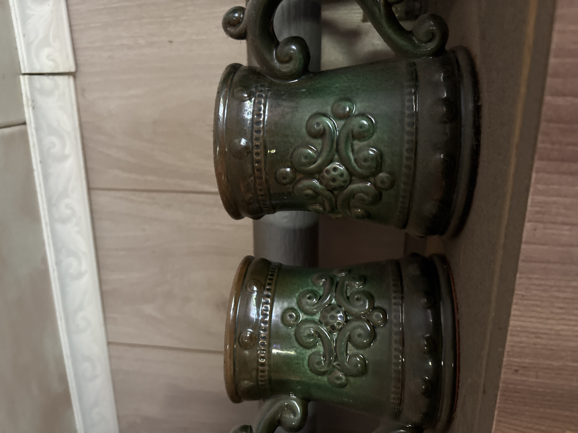 Two dark green, decorative ceramic mugs with handles and embossed swirling patterns are displayed on a wooden shelf. The mugs have a glossy finish and a rim of a lighter brown color. Behind the mugs, a light brown tiled wall is visible, topped by a white decorative border. The shelf is made of wood with visible grain. The scene appears to be indoors, possibly in a home or shop. The image is a close-up shot, focusing on the mugs and their immediate surroundings.