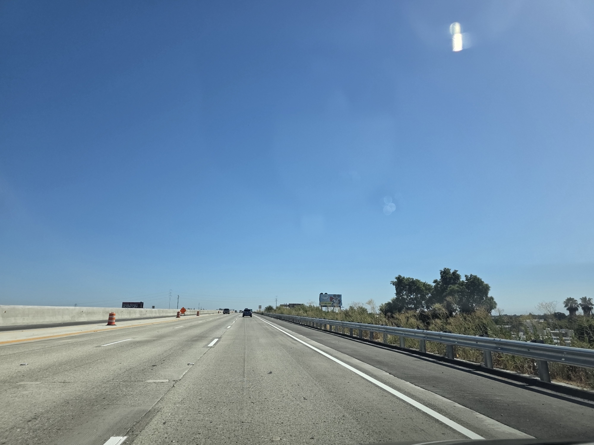 The media file depicts a view from a vehicle traveling on a highway under a clear, bright blue sky. The highway has multiple lanes, and traffic appears light with a few cars visible in the distance. Orange traffic cones are placed along the side of the road, indicating construction or a lane closure. A concrete barrier separates the highway lanes from an adjacent area.
To the right of the highway, there is a metal guardrail bordering a vegetated area with dry grasses and some trees. In the distance, a billboard can be seen with colorful graphics and text, though the specifics are illegible. The time of day appears to be midday, given the bright sunlight and lack of shadows. The weather is clear and sunny.
The scene suggests a typical highway driving experience on a sunny day in a potentially rural or suburban area, given the roadside vegetation and open sky. There is no discernible emotion conveyed. The location is identified as West Sacramento, United States of America.