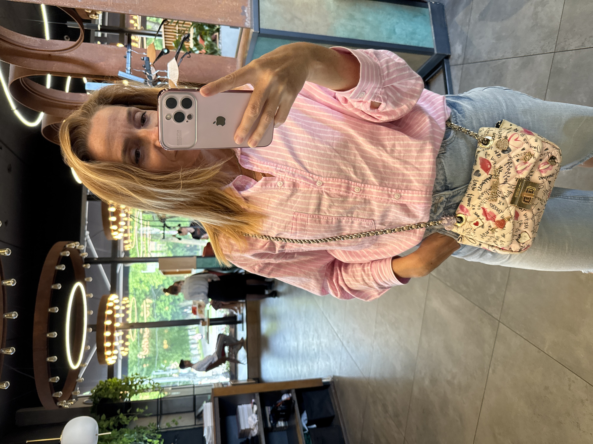 A woman takes a selfie in a cafe in Veliky Novgorod, Russia. She is wearing a pink and white striped button-down shirt and light blue jeans. She is holding a pink smartphone in her right hand and a small, quilted handbag with a chain strap is visible on her left hip. The handbag is decorated with pink hearts, red lips, and script text. The background shows a modern cafe interior with large windows, plants, and industrial-style light fixtures. Several other patrons are visible in the background, including a man sitting at a table and a woman standing. The floor is tiled.