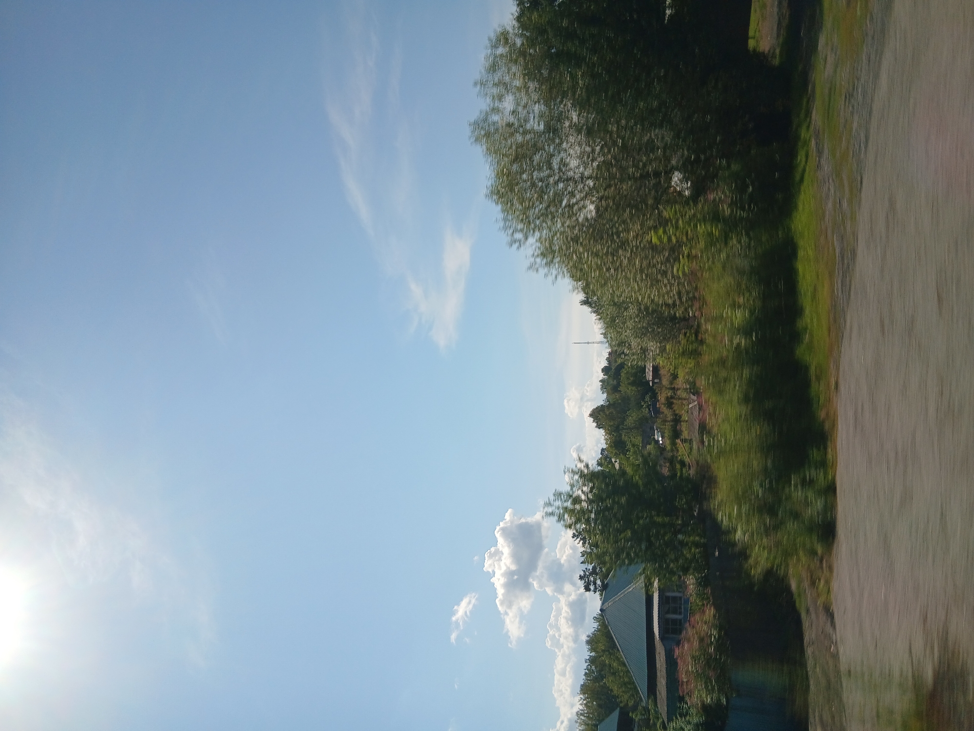 The image shows a view of a rural area on a sunny day. The sky is clear blue with a few white clouds and the sun is visible in the upper left corner, casting a bright light. In the middle ground, there are several houses with green roofs, surrounded by trees and vegetation. Some of the trees are in bloom, with pink and white flowers. The foreground consists of a dirt road or path. The overall scene is peaceful and depicts a natural, undeveloped landscape.