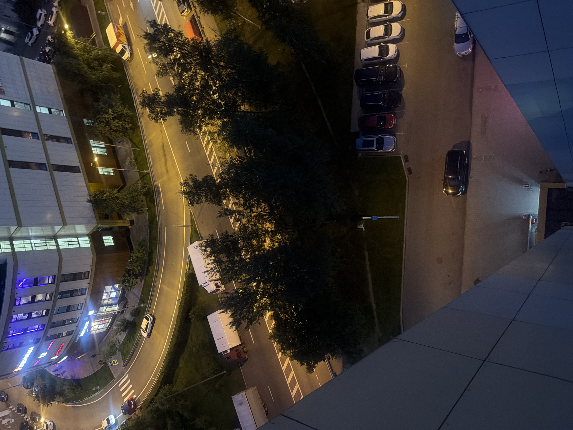 An aerial shot captures a street scene at dusk in the city of Dyuna SSK, Russia, with a modern building dominating the left side. The building, illuminated with neon blue lights, features a curved facade and several stories of windows. A road curves around the building, with several vehicles visible. A white SUV drives on the road, and a red car follows behind. Further down the road, two white box trucks are parked, and a red truck is also visible. A white delivery truck with a red and yellow logo drives by. Across the street, a row of parked cars, including a black SUV and several white sedans, are visible in a parking lot. Trees line the street and parking lot, adding a touch of nature to the urban landscape. The overall impression is one of a bustling city at night, with a mix of modern architecture and natural elements.
