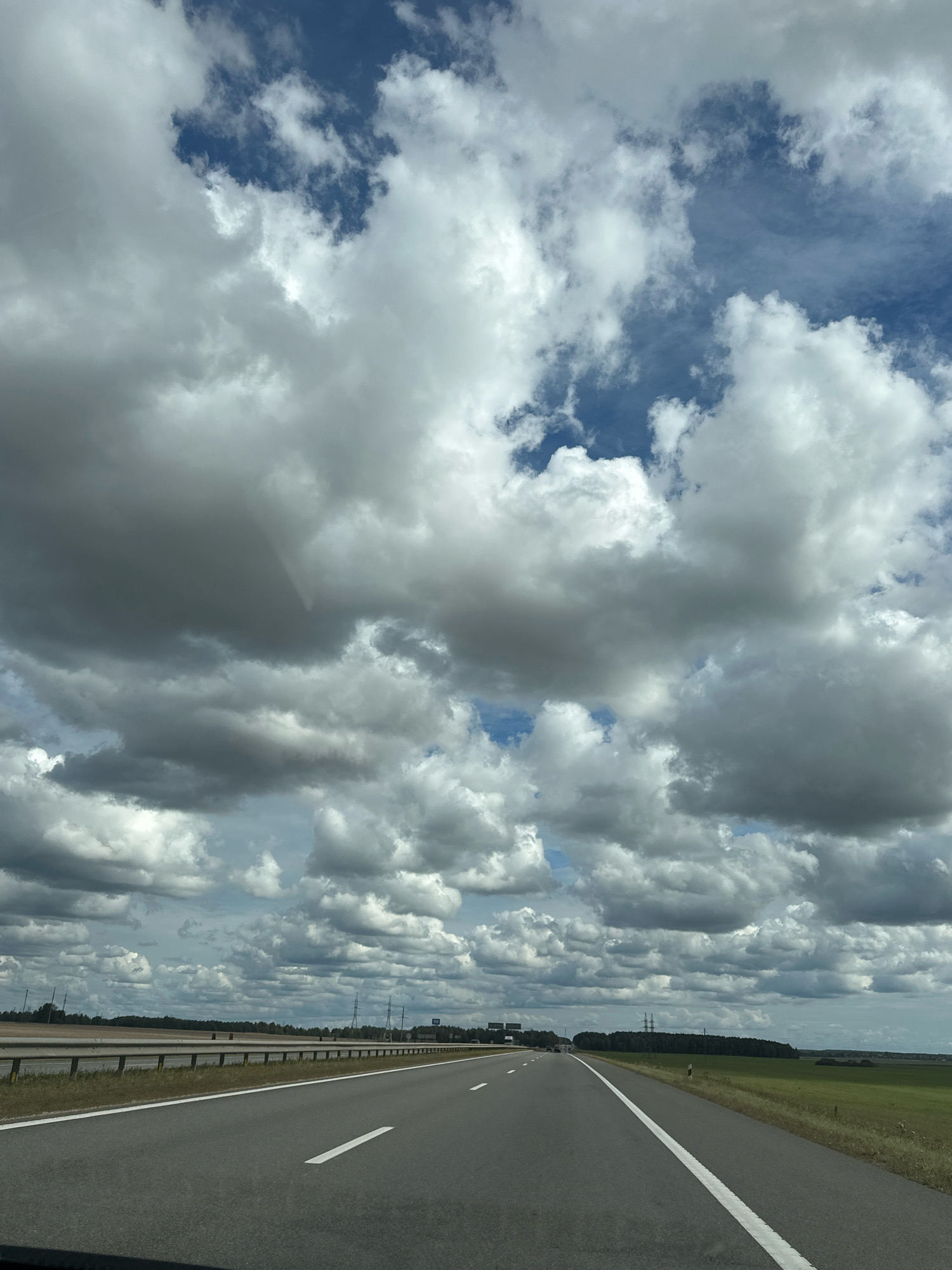 The image shows a view from a vehicle driving on a highway. The highway is paved and has white lane markings. There is a metal guardrail on the left side of the road, separating the highway from a grassy area. In the distance, there is a treeline and what appears to be agricultural fields. The sky is filled with dramatic, large clouds, with patches of blue sky visible in between. The overall scene suggests a rural or semi-rural environment in Belarus, likely on a cloudy day.