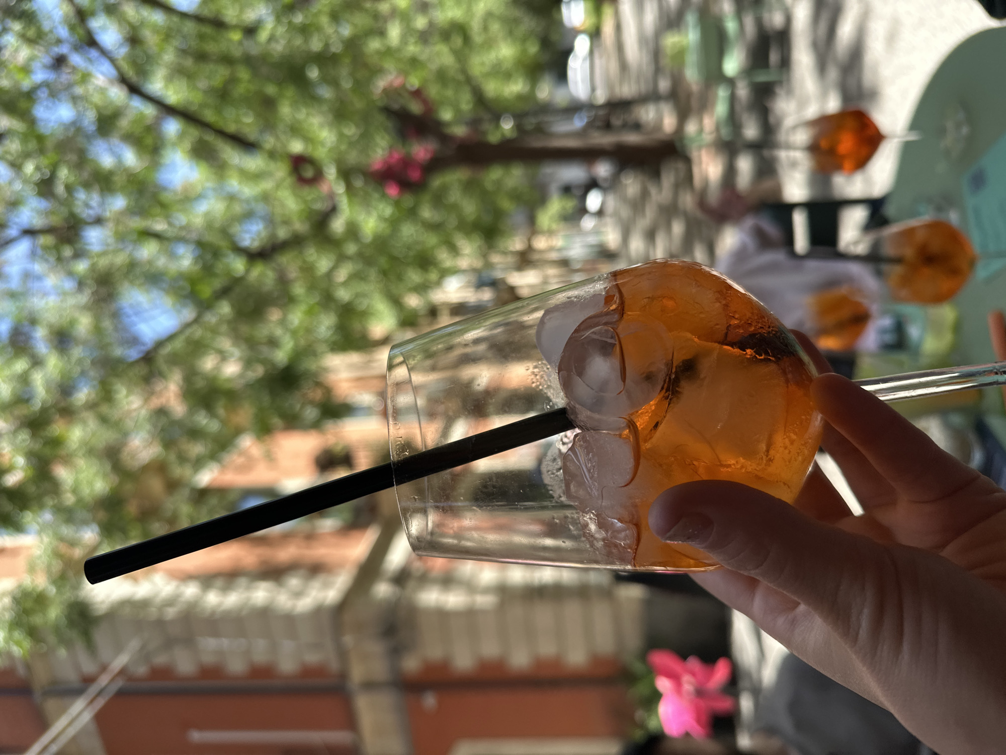A close-up, slightly blurred, rotated eye-level shot shows a person holding a glass with an orange drink and ice. A black straw is inside the glass. Another straw, seemingly glass, is outside the glass. Other similar glasses can be seen on a nearby table. The background is a mix of trees and buildings, with a reddish-brown building facade partly visible. The location is likely in Catania, Italy.