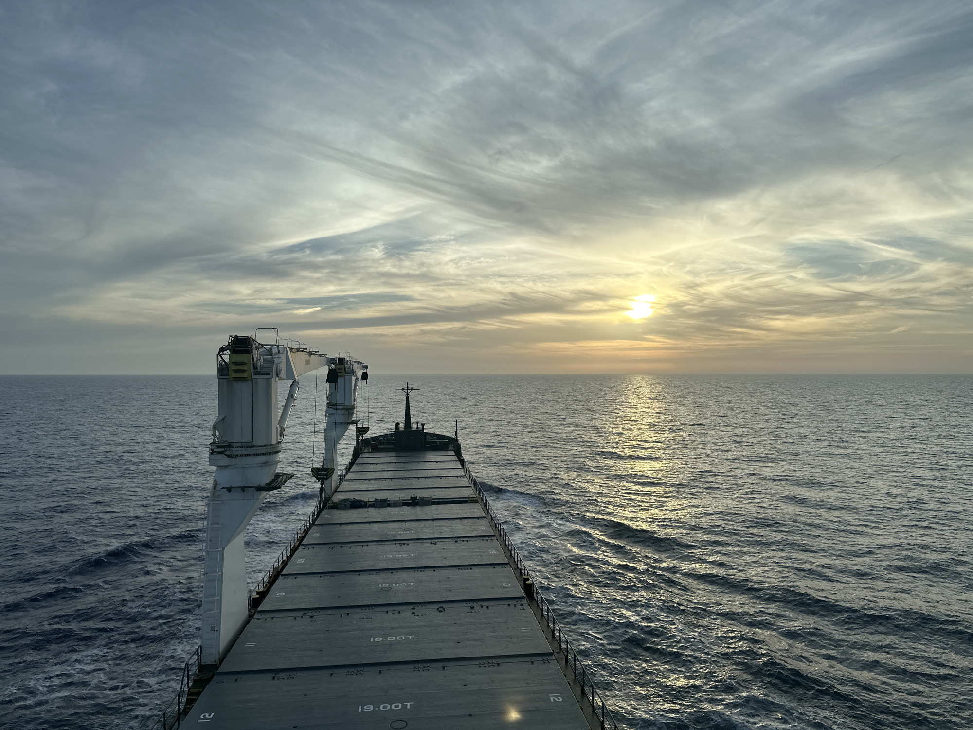 The image shows the deck of a large cargo ship sailing on the open ocean at sunset. A crane stands on the left side of the ship. The deck consists of large, flat panels, some of which are labeled with numbers and "19.00T". The ocean stretches to the horizon, reflecting the golden light of the setting sun. The sky is filled with streaky clouds. The location is the open ocean, not a city.