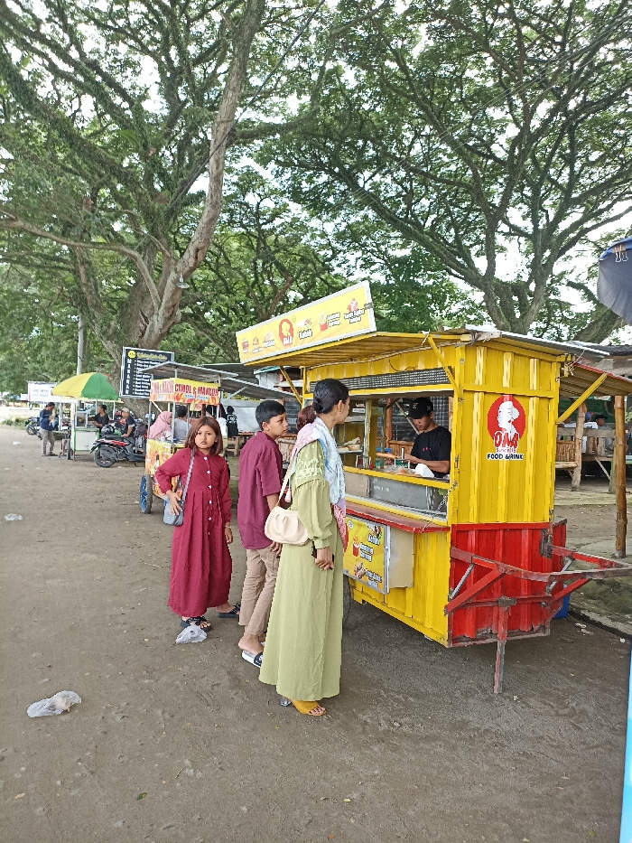 This daytime image captures a vibrant street food scene in Kepung, Indonesia, set beneath the expansive canopy of large, mature trees. The ground is a mixture of dirt and gravel, indicating an informal outdoor setting.
In the foreground, a family group stands near a brightly colored food stall. A woman, dressed in a long light-green garment and a headscarf, carries a beige handbag and stands facing the stall. To her left, a boy in a maroon top and light trousers stands looking slightly away. Beside him, a young girl in a long red dress looks directly at the camera, holding a small bag.
The most prominent object is a mobile food cart, painted yellow with red accents at its base and wheels. Inside, a vendor in a black shirt and cap is visible, preparing food or drinks. The cart features several signs: a large circular logo with a chef's hat and the text "OMA FOOD DRINK," along with a smaller rectangular sign showing images of food like fries and burgers, and the text "FOOD GRILLED." Above the serving window, another banner displays various food icons and partially legible text.
Further in the background, another yellow food cart is visible, with a banner advertising "MIE AYAM CHOCO & MANTEP" and "COOL & MANTEP," suggesting chicken noodles and other items. Several motorcycles are parked nearby, and more people are scattered around, indicating a lively local gathering spot. The overall impression is one of casual outdoor dining and community activity. The lighting is diffused, suggesting an overcast day or shaded conditions. A few discarded plastic bags are visible on the ground.