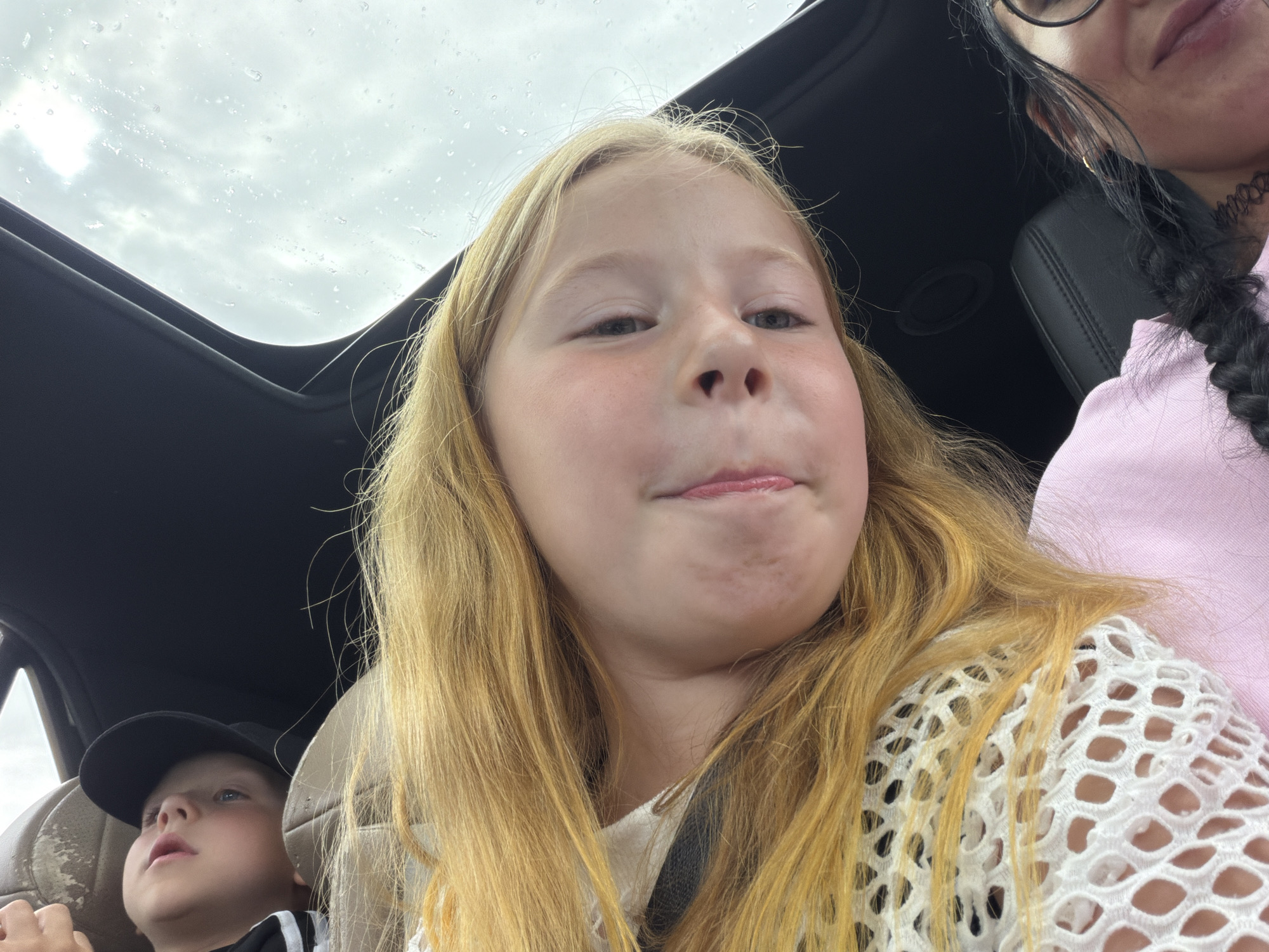 In the image, a young girl with long blonde hair is taking a selfie. She is in the front passenger seat of a car, with a sunroof open above her. The sky is overcast and appears to be raining, with visible water droplets on the sunroof. To her left, a young boy wearing a black baseball cap and a black and white striped shirt is visible in a car seat. To the right, a portion of an adult's face with dark hair and glasses can be seen. The adult is wearing a pink shirt and a black choker necklace. The girl is wearing a white mesh top over a white shirt. The overall mood of the image is casual and appears to capture a moment during a car ride. There is no visible text in the image. The location context is Шолохово, Russia.