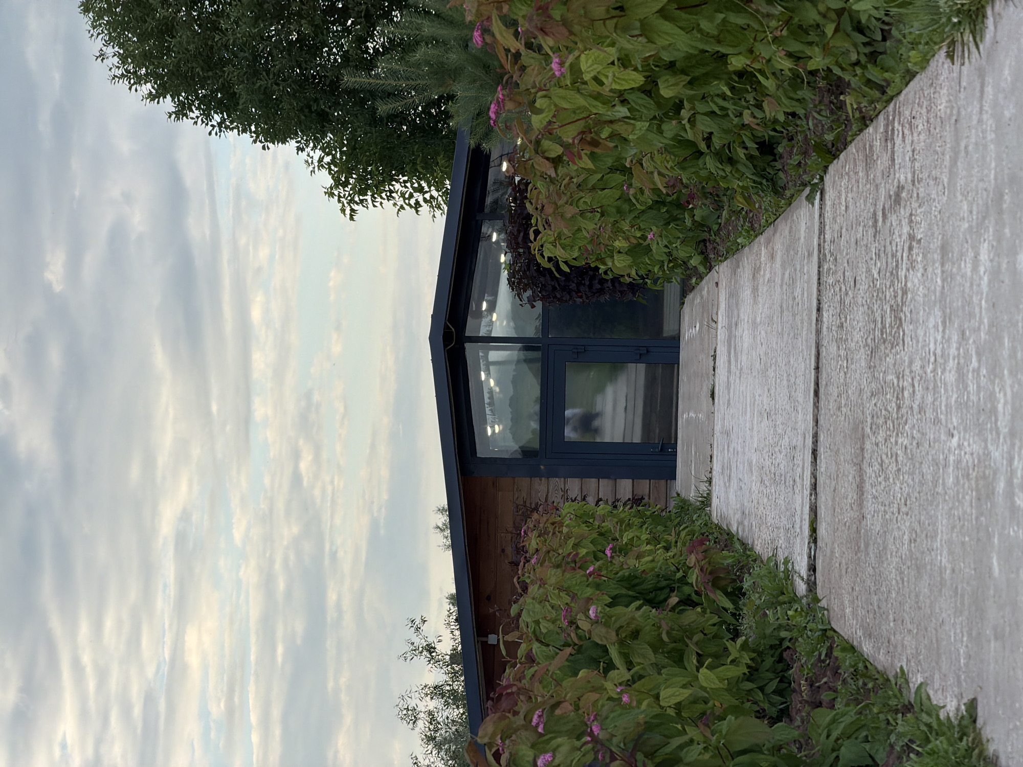 A concrete walkway leads to a modern building with a wooden exterior and a glass facade in Bol'shaya Ven'ya, Russia. The walkway is flanked by lush green bushes with small pink flowers. The sky above is overcast with a mix of gray and white clouds. The building's glass walls reflect the interior lighting and a blurred image of a person walking. A tall green tree stands to the right of the building.