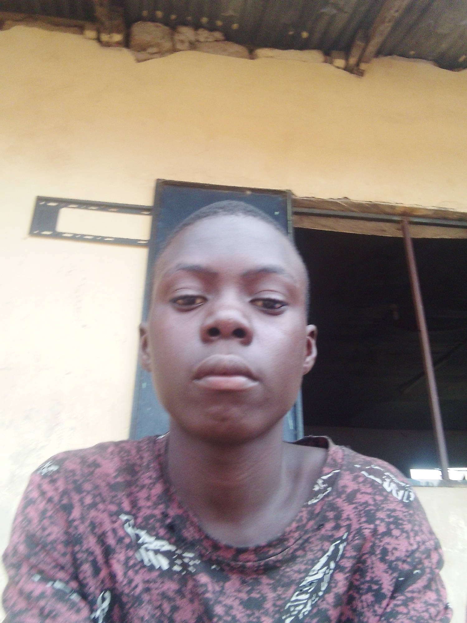 A person is standing in front of a yellow wall with a window. The person has dark skin, short hair, and is wearing a patterned shirt. The wall is textured and has an object mounted on it. The window is dark and has a bar visible. The location is Unknown, Unknown.