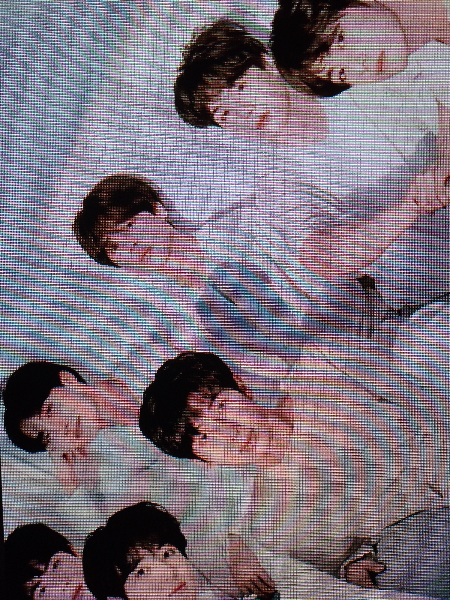 A group of seven young men, seemingly East Asian, are posed closely together. They are predominantly dressed in light-colored tops, possibly white t-shirts. They appear to be lying down, possibly on a bed, although the exact environment is obscured. Their expressions are relaxed, some smiling, and they are looking towards the camera. The image quality is pixelated, which suggests it's a close-up of a digital display. There is no further detail known about location or setting.