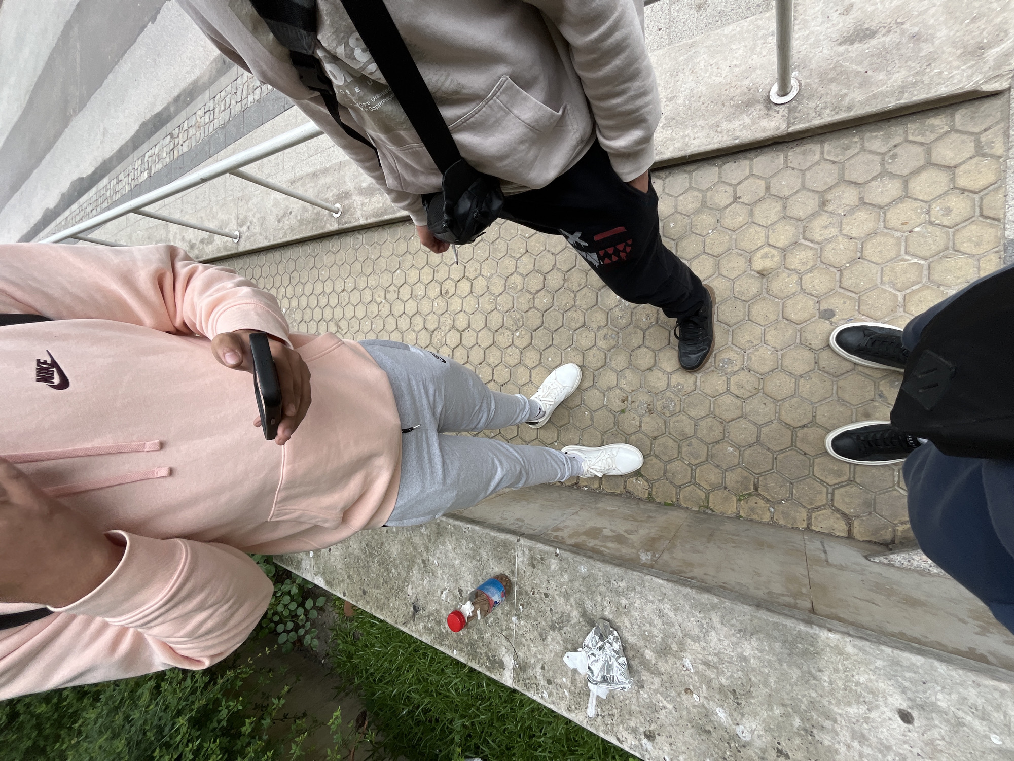 The image, taken from an elevated viewpoint, shows three individuals standing on a honeycomb-patterned pavement next to a concrete ledge with foliage below. One person wears a light pink Nike hoodie and grey sweatpants with white sneakers, holding a black phone. Another wears a light beige hoodie, black sweatpants, and black sneakers with a black cross-body bag. A third individual is partially visible, wearing navy blue pants and black sneakers. On the concrete ledge are litter items: a plastic bottle and a foil wrapper. The location appears to be near a metal railing with grey steps behind it. The location is likely in Plovdiv, Bulgaria.