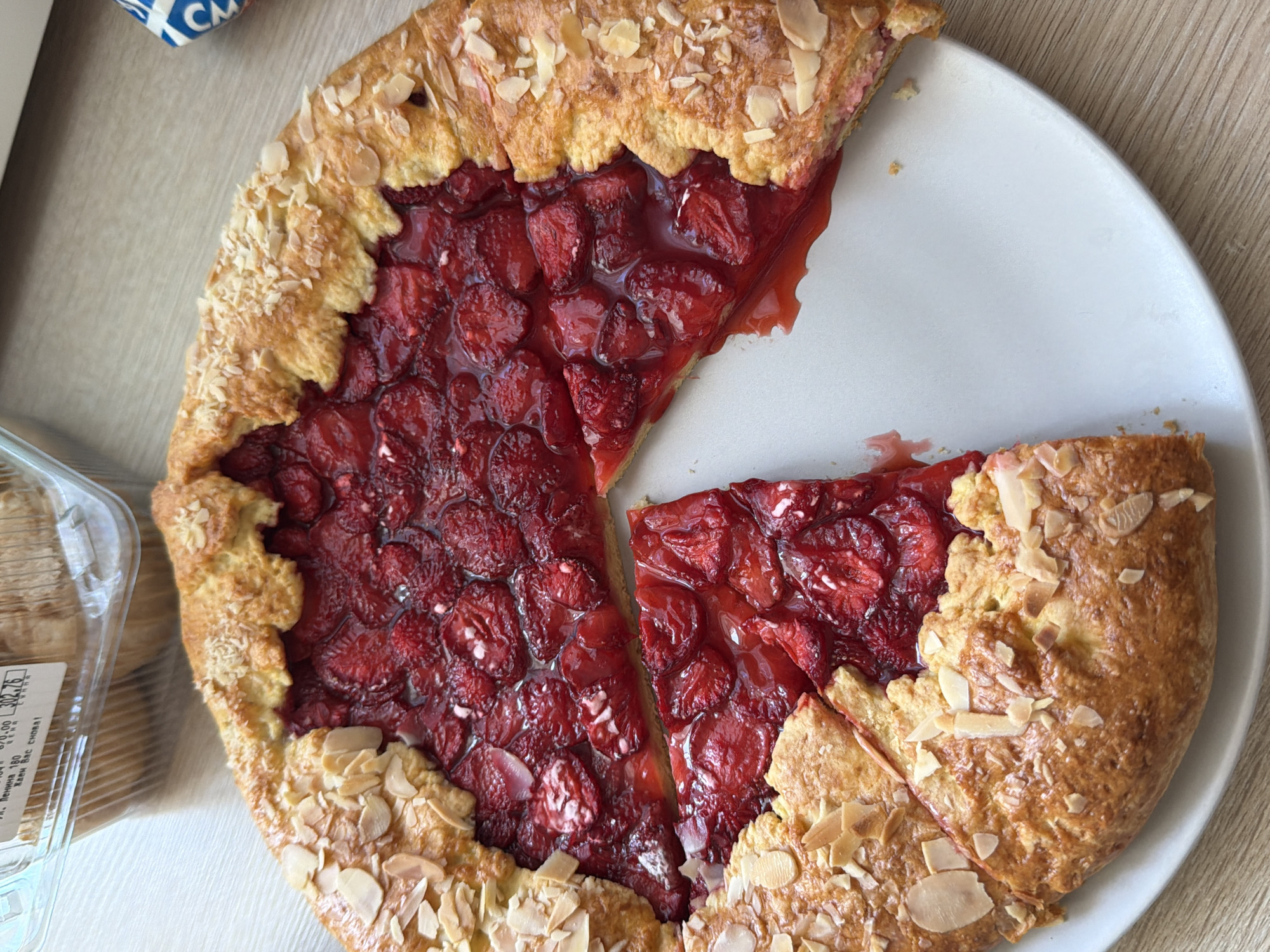 A partially eaten strawberry galette sits on a white plate on a wooden table. The galette is round with a flaky golden-brown crust, sprinkled with sliced almonds. The filling consists of sliced strawberries in a glossy, red syrup. One slice has been removed from the galette and placed next to it on the plate. In the upper left corner of the image, a clear plastic container with a label is visible, indicating it likely held the galette. The label is partially obscured but shows Cyrillic text and numerical information. The scene is indoors, with the lighting suggesting it is daytime. There are no people or animals visible in the image. The environment appears to be a domestic setting, likely a kitchen or dining area. The context of Izhevsk, Russia is provided but cannot be visually confirmed from the image alone.