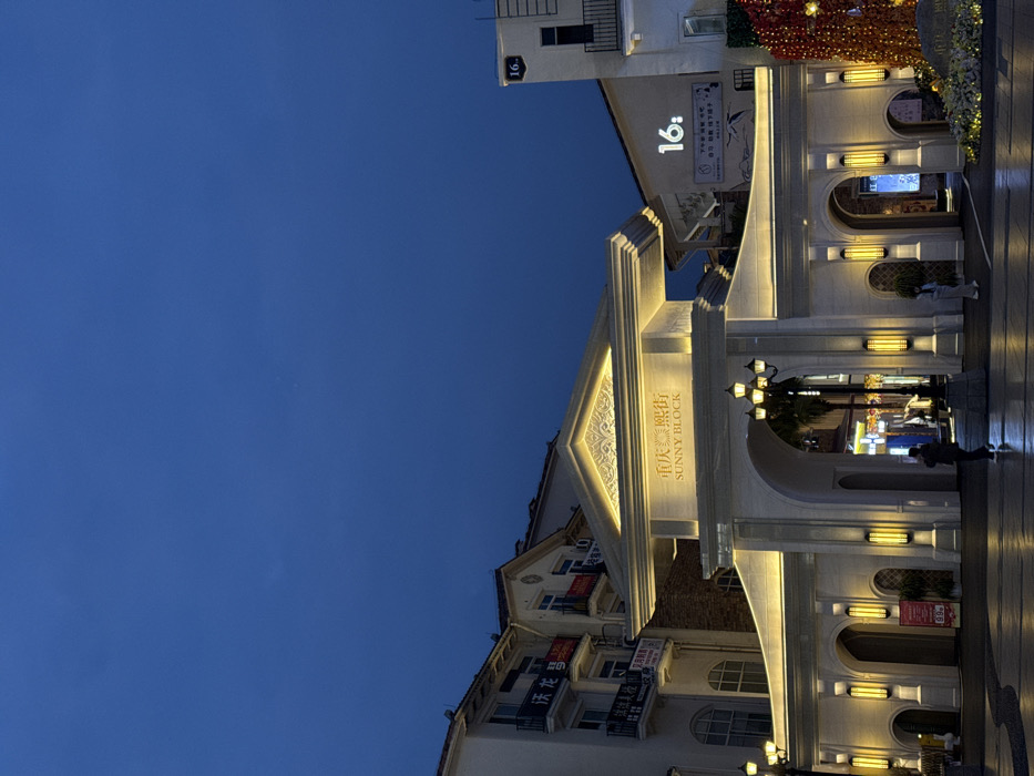 The image displays an illuminated outdoor scene at night within the Chongqing High-tech Zone, China, focusing on an ornate entrance structure. A large, light-colored archway, designed in a classical European style, dominates the center of the frame, featuring the illuminated text "重庆 熙街 SUNNY BLOCK" at its apex. The archway is brightly lit by warm white LED strips along its cornices and vertical sconces on its pillars.
To the left of the archway, a multi-story building with balconies and traditional architecture is visible, displaying banners with Chinese characters. To the right, another multi-story building, similarly styled, features the illuminated number "16:" on its facade. The foreground consists of a patterned, dark-paved plaza, illuminated by multiple street lamps with triple lantern heads.
Two individuals are present in the plaza: one person, dressed in dark clothing, walks centrally towards the archway's illuminated interior, and another person is partially visible further to the right. On the far right, decorative elements include dense clusters of warm-toned string lights, possibly red or orange, draped over a structure or foliage, along with what appears to be a flower arrangement at ground level. The sky above is a clear, deep blue, consistent with twilight or nighttime conditions.