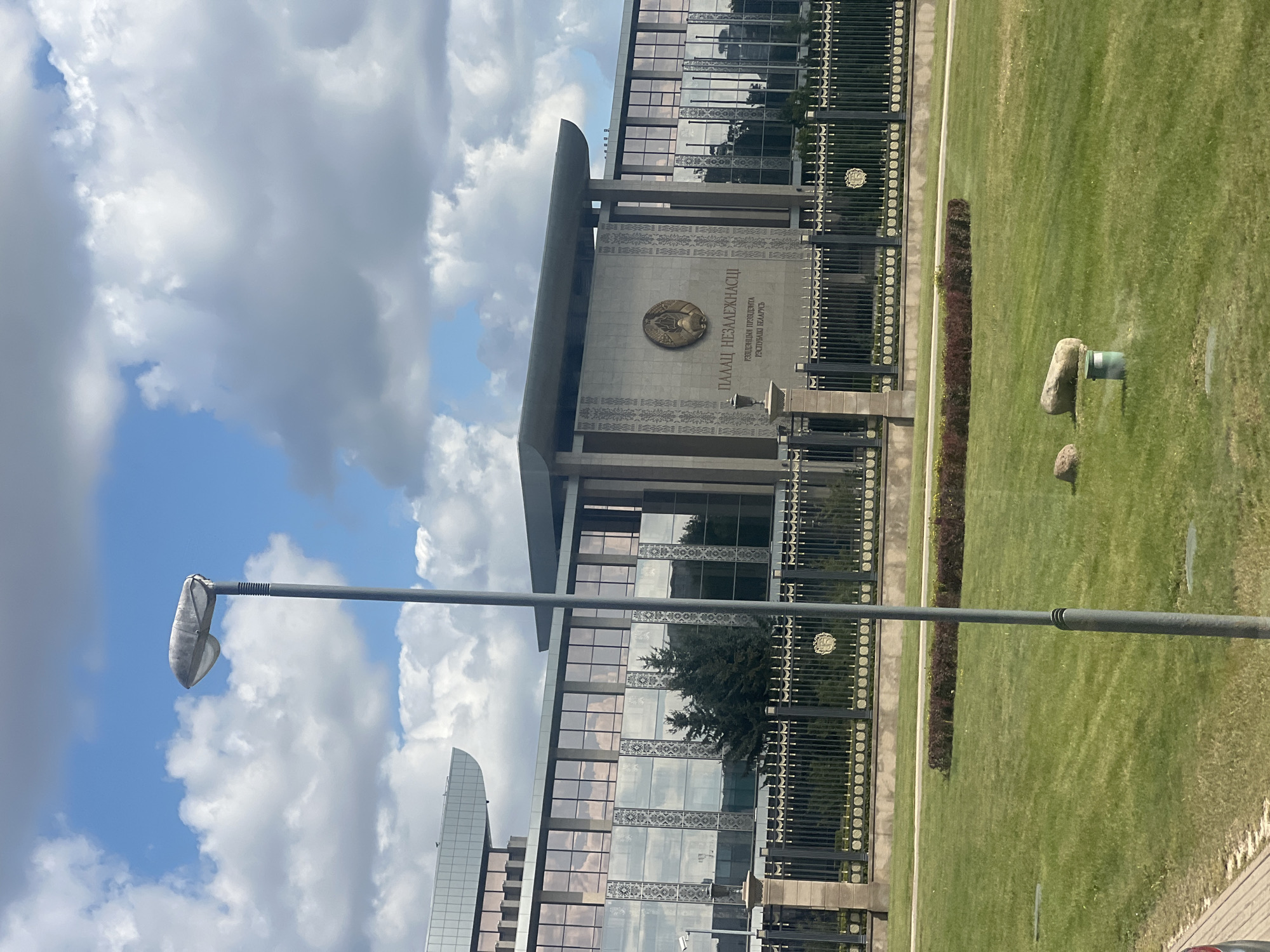 The image shows the Palace of Independence in Minsk, Belarus, on a partly cloudy day. The modern building features large glass windows and a central facade with Cyrillic text and a coat of arms. A tall, grey lamppost stands in the foreground, to the left of the center. In front of the building, a black metal fence with decorative elements encloses a manicured lawn. Beyond the fence, a strip of dark red bushes runs parallel to the fence. The foreground consists of a well-maintained green lawn with a few small rocks and a sprinkler head visible. The sky is a bright blue with fluffy white clouds. There are no people visible in the image.