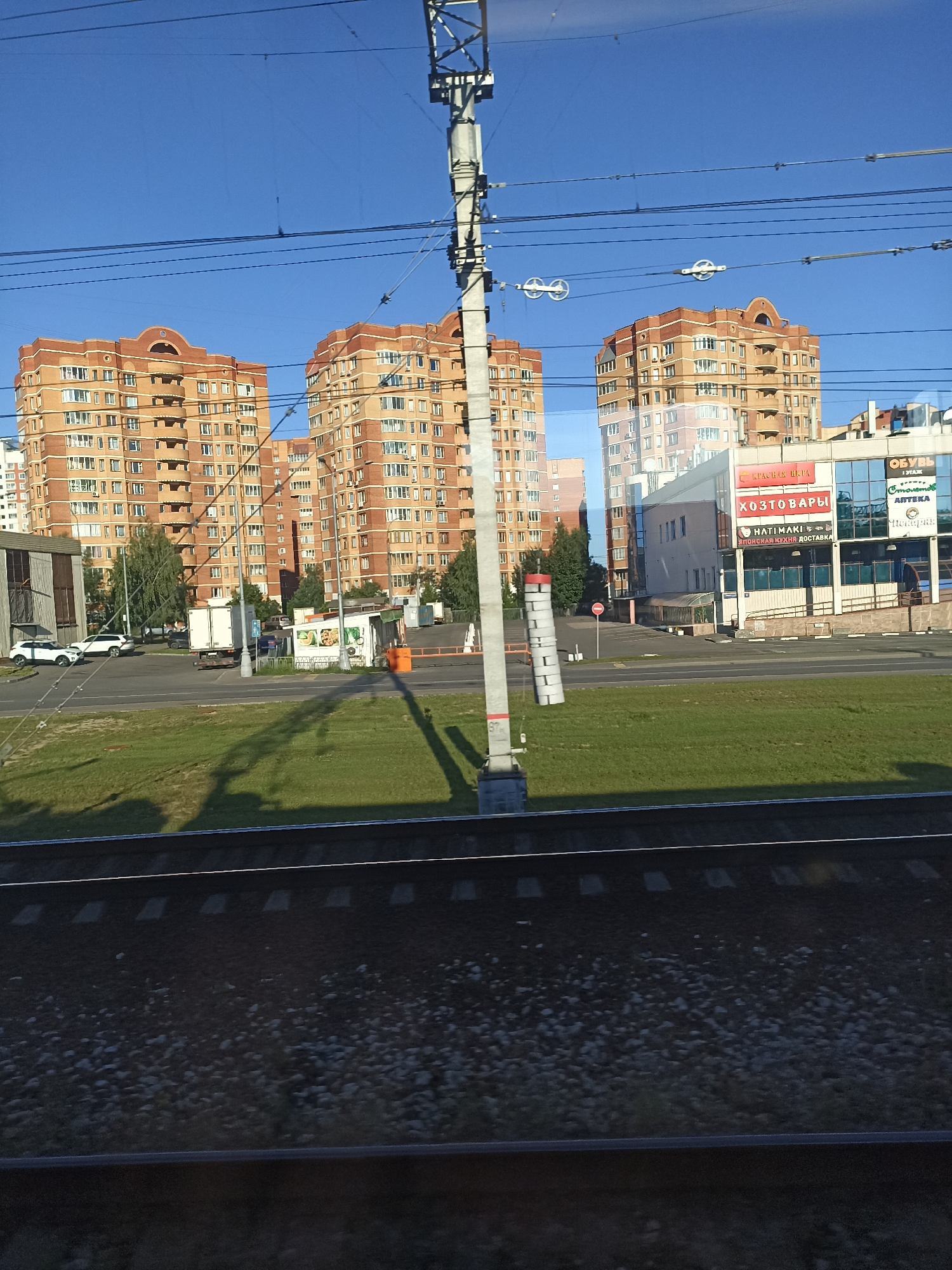A low-angle shot from a train captures a sunlit urban scene in Shcherbinka, Russia, dominated by several large, salmon-colored apartment buildings with arched rooflines. In the foreground, train tracks stretch horizontally across the frame, with ballast and ties visible. Beyond the tracks lies a strip of manicured green grass, casting long shadows from an unseen pole and overhead wires.
A paved road runs parallel to the grass, with several vehicles present: two white SUVs, a white box truck, and a dark-colored passenger car. Adjacent to the road is a small, white, kiosk-like structure with colorful graphics depicting food items, possibly a food vendor. Further back, a modern commercial building with large glass windows displays several signs in Russian, including "КРАСНАЯ ЦЕНА" (Red Price), "ХОЗТОВАРЫ" (Household Goods), "HATIMAKI" and "ЯПОНСКАЯ КУХНЯ ДОСТАВКА" (Japanese Cuisine Delivery), and "ОБУВЬ" (Shoes).
Overhead, a complex network of electrical wires and support structures for a trolleybus or tram system crisscrosses the bright blue sky. The overall impression is of a typical day in a Russian residential and commercial district. No people are clearly visible in the image.