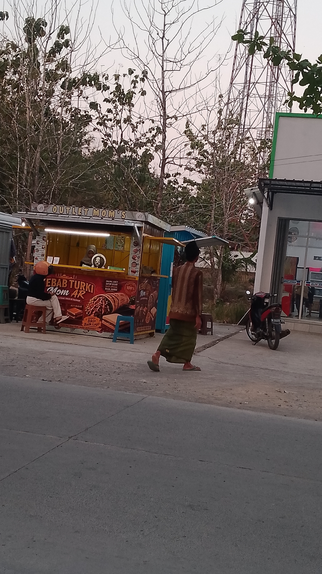 The image depicts an outdoor scene at dusk, likely in Karanglangu, Indonesia. On the left, a food stall named "OUTLET MOM'S" with yellow and blue accents sells "KEBAB TURKI". A person wearing a helmet sits on a stool in front of the stall. To the right of the stall, a person wearing a traditional patterned top and a green sarong walks away from the camera. A red motorcycle is parked near a building with a green and white facade on the right side of the image. Bare trees and a tall metal tower dominate the background. The ground is paved with asphalt and dirt.