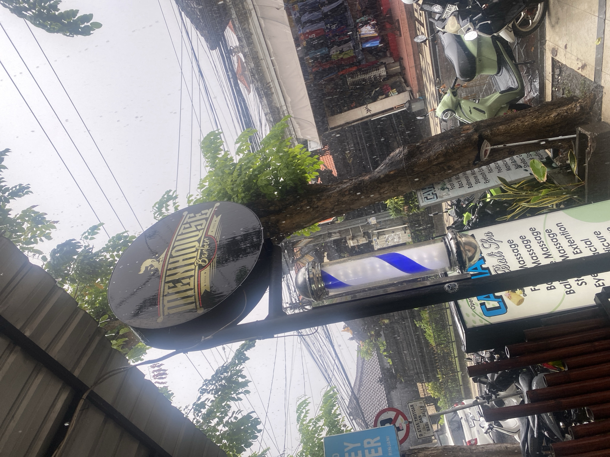 The image captures a street scene in Kuta, Indonesia, showing a "MEHHOUSE Barber" sign alongside a barber pole. A spa sign is also visible. A scooter is parked nearby, and the street appears to be wet, suggesting recent rain. Utility lines crisscross overhead. A street sign is partially visible at the bottom.