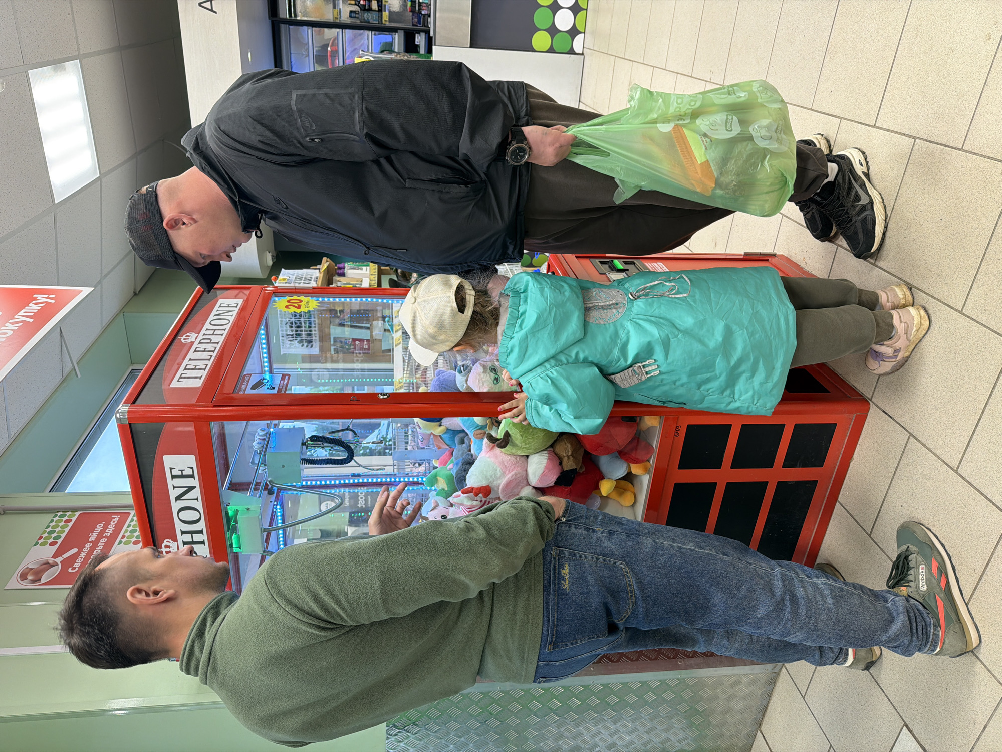 A father wearing a black jacket and black cap stands next to a young child in a turquoise jacket and white cap as they look into a red claw machine with plush toys. Another man in a green sweater and jeans is beside the claw machine. The location appears to be a retail establishment in Chaikovsky, Russia, based on signage in the background. The father holds a green plastic bag. The child's hands are resting on the glass of the machine, appearing to examine the toys.