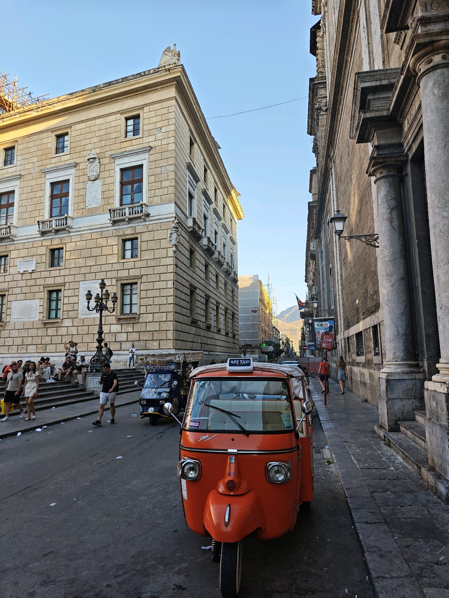 In Palermo, Italy, an orange Ape taxi is parked on a street lined with tall, historic buildings. The taxi is a three-wheeled vehicle with a small cabin and an open rear. The driver's cabin is visible, with a sign that reads "APE TAXI" on the roof. Beside the orange taxi, a blue Ape taxi is also visible. Several people are walking on the sidewalk and sitting on the steps of a building. The street is paved with cobblestones, and there are some scattered pieces of litter. The buildings are made of stone and have a classical architectural style. The sky is clear and blue. The image appears to have been taken in the late afternoon or early evening, as the sun is casting long shadows.