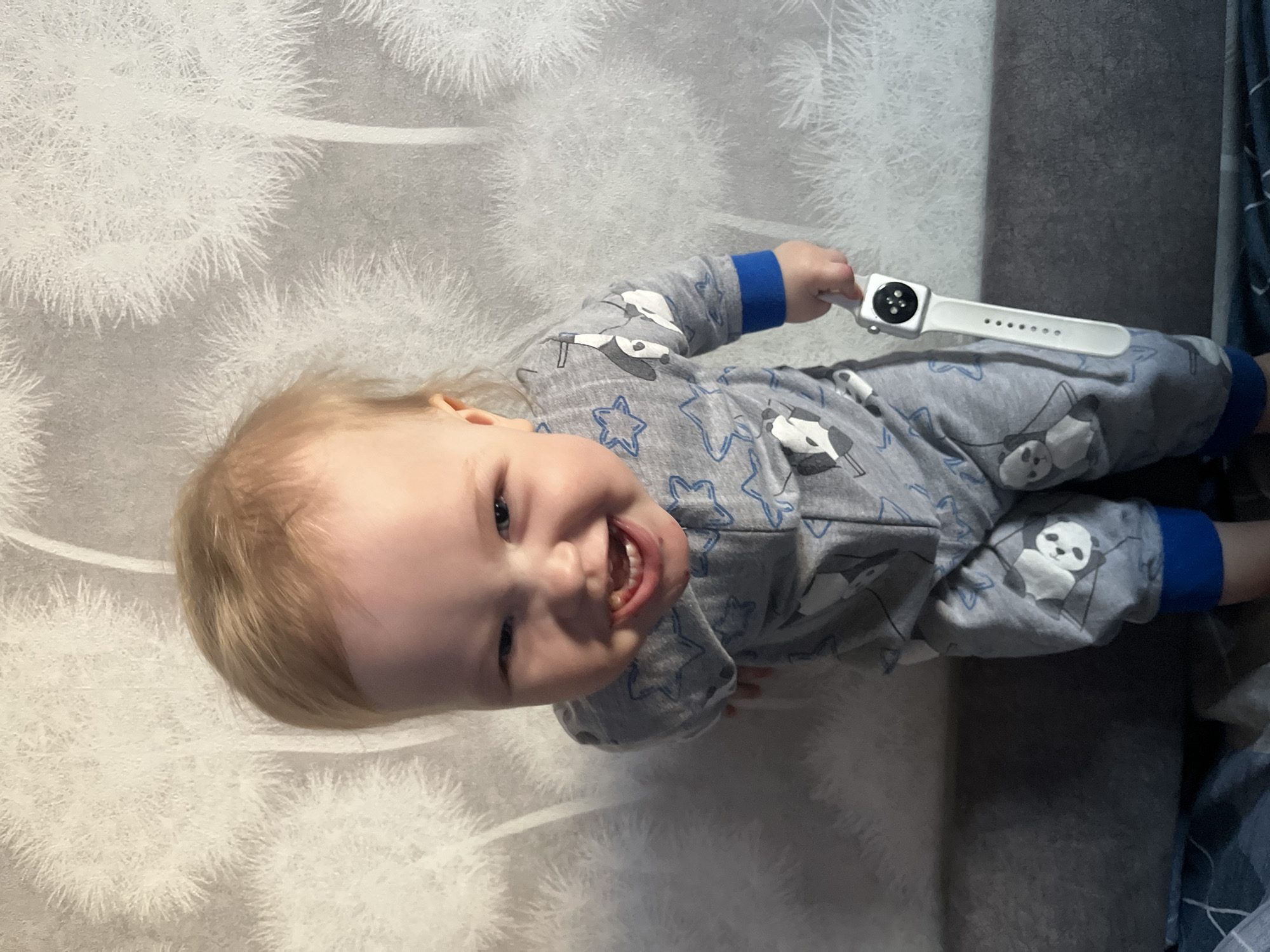 A baby lies on a patterned white and gray blanket. The baby is wearing a gray pajama set with blue cuffs, a star pattern, and panda bear graphics. The baby holds a white smartwatch in their right hand. The location may be Chaikovsky, Russia, but the evidence is not present in the image.