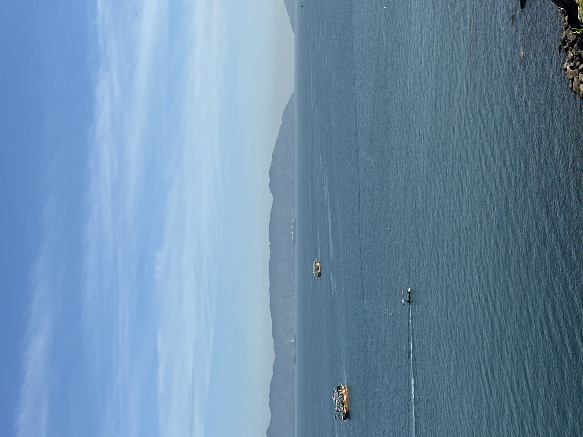 A wide shot captures the calm blue waters of the sea under a clear sky with wispy clouds. In the distance, a hazy mountain range stretches across the horizon. Several boats are visible: a larger, orange-hulled vessel with a canopy is moored on the left, while a smaller, green-roofed boat is further out. A white boat with a red flag is speeding across the water, leaving a wake. The foreground shows a rocky shoreline. The scene suggests a view of the coastline near Da Nang, Vietnam, potentially from a vantage point overlooking the South China Sea.