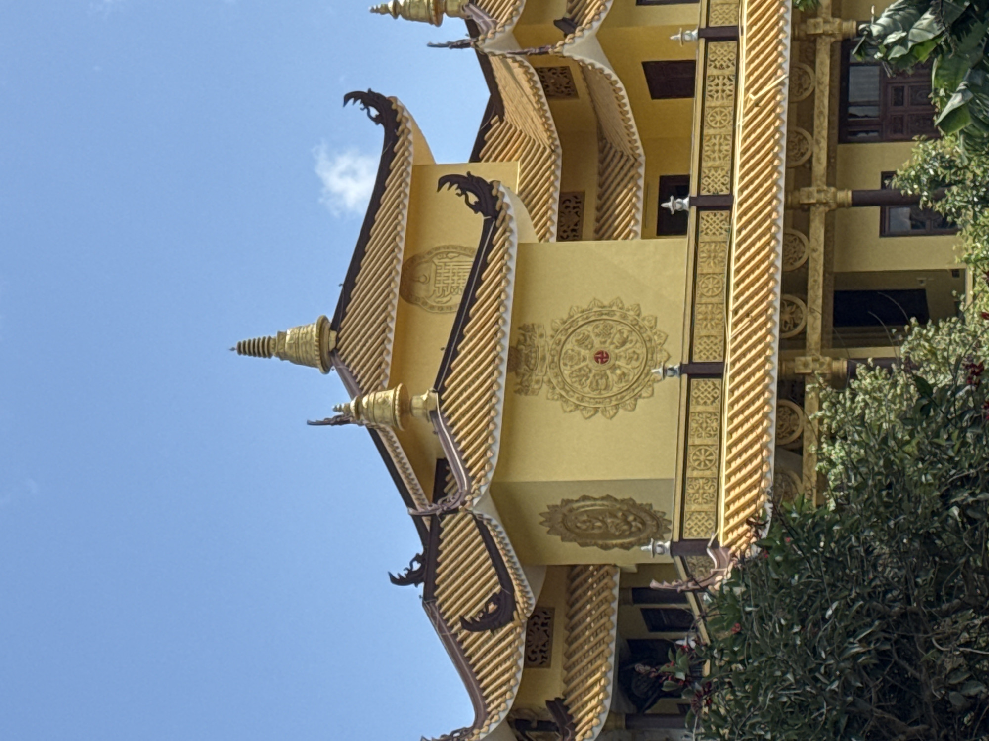 The image shows the exterior of a vibrant yellow Buddhist temple with golden, tiered roofs and ornate carvings. The temple features several prominent architectural elements, including curved eaves, finials, and decorative circular motifs on its walls. Lush green foliage, likely trees and bushes, partially obscures the lower section of the building, suggesting an outdoor setting with natural landscaping. The sky above is a clear, bright blue with a few wispy clouds. This temple is identified as the Truc Lam Zen Monastery, located on Phuong Hoang Mountain overlooking Tuyen Lam Lake in Da Lat, Vietnam.