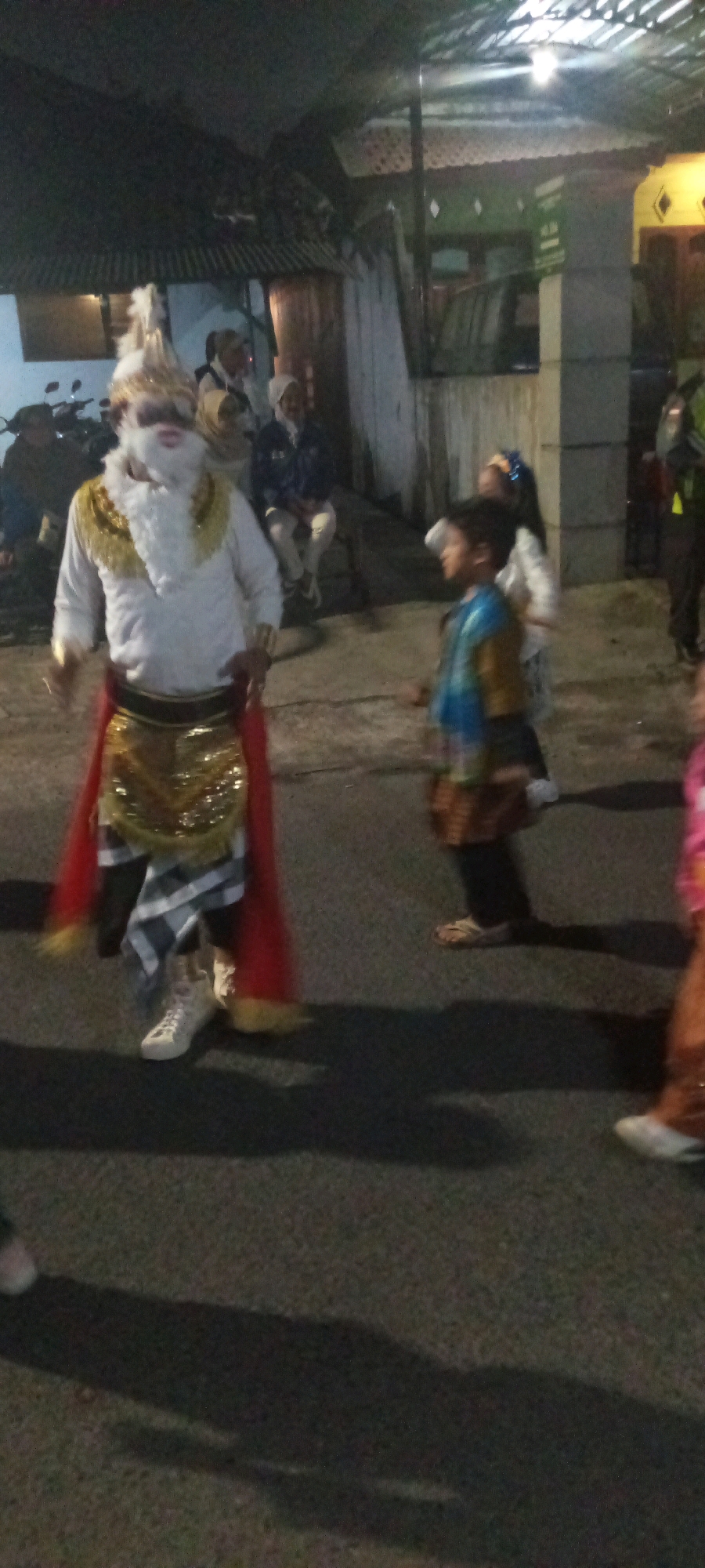 In an outdoor setting, likely a street or pathway at night, a group of people are visible. The central figure is a person in elaborate costume, featuring a white shirt with gold trim around the shoulders, a sparkling gold sash at the waist, and a red cape draped around their legs. They are also wearing a patterned skirt and white sneakers. This costumed individual appears to have a white beard and a festive headpiece. To the right of this person, a young boy in a blue and gold outfit is looking to his left, with a basket-like object in front of him. Behind the central figure, several other individuals are present, including two women in headscarves and one person in a dark jacket and lighter pants. In the background, buildings with corrugated roofs and illuminated windows are visible, along with a parked motorcycle on the left. The ground is paved, and shadows are cast on it, suggesting artificial lighting. The overall scene depicts a festive gathering or performance.