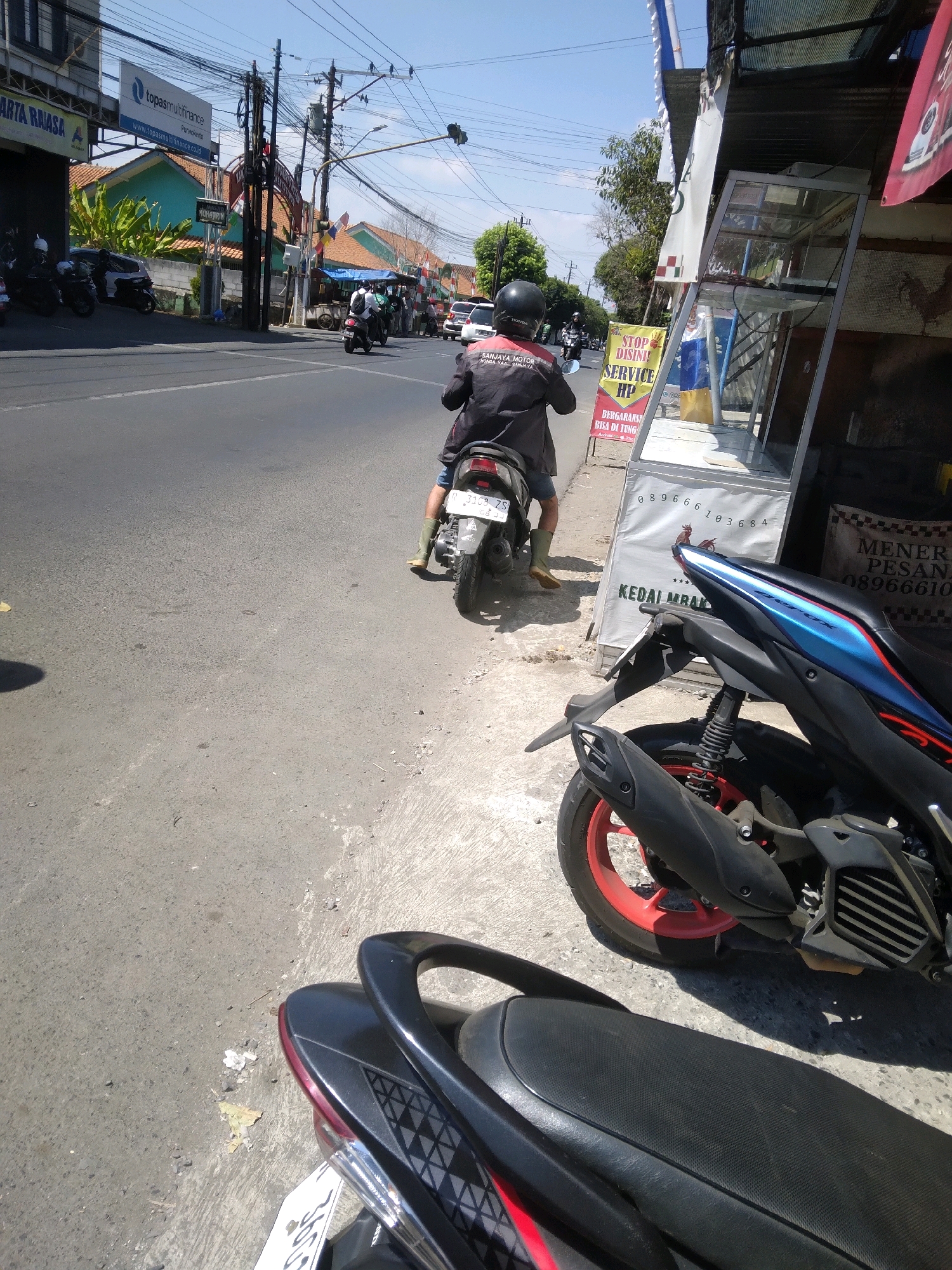 The image shows a street scene in Purwokerto, Indonesia. In the foreground, a person is riding a motorcycle. The rider is wearing a black jacket with white lettering on the back. In the background, there are other motorcycles, cars, and buildings. There is also a sign that reads "Topas Multifiance Purwokerto." The sky is clear and blue, and the sun is shining.