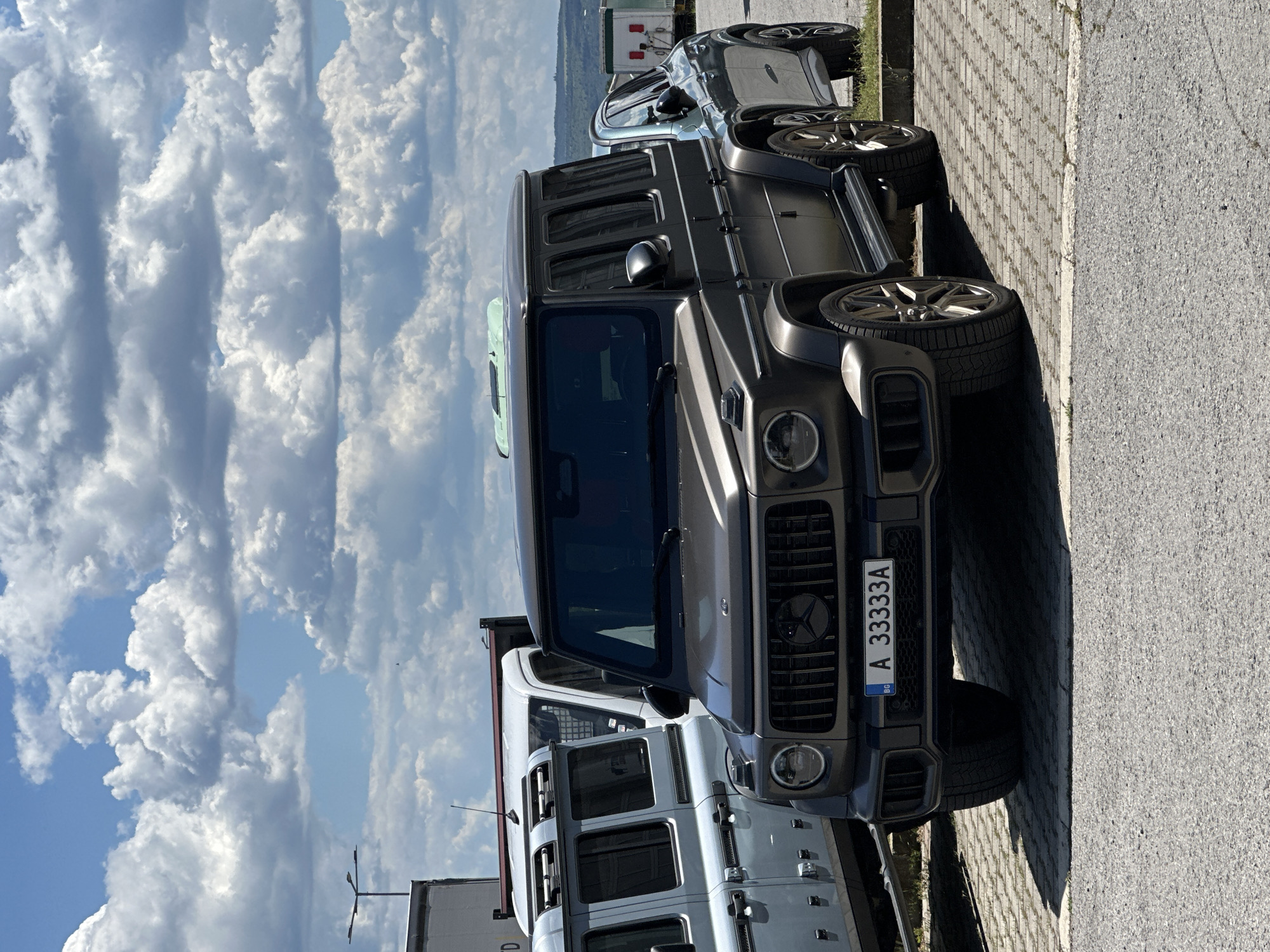 The image shows several cars parked in Varna, Bulgaria. The prominent car is a dark gray Mercedes-Benz G-Class with a Bulgarian license plate "A 33333A". Beside it is a white car, potentially a Land Rover Defender, with a partial view. Further back, another car is visible. The vehicles are parked on a paved surface. The sky is cloudy with some blue patches visible. In the distance, some buildings and greenery can be seen. No people are visible in the image. The orientation of the image is rotated 90 degrees counter-clockwise.