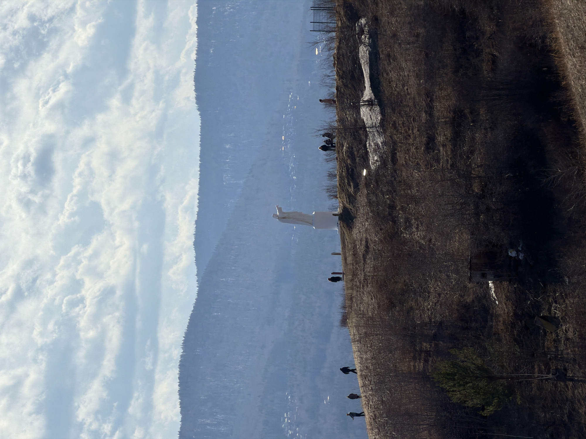 The image shows the landmark "Tsar Fish" monument located in Krasnoyarsk, Russia, perched on a hillside overlooking the Yenisey River. The sky is filled with white clouds, the mountain side consists of brown shrubs, trees, and tall grass. In the center of the scene is the white statue of the Tsar Fish monument. There are several people standing along the hillside.
