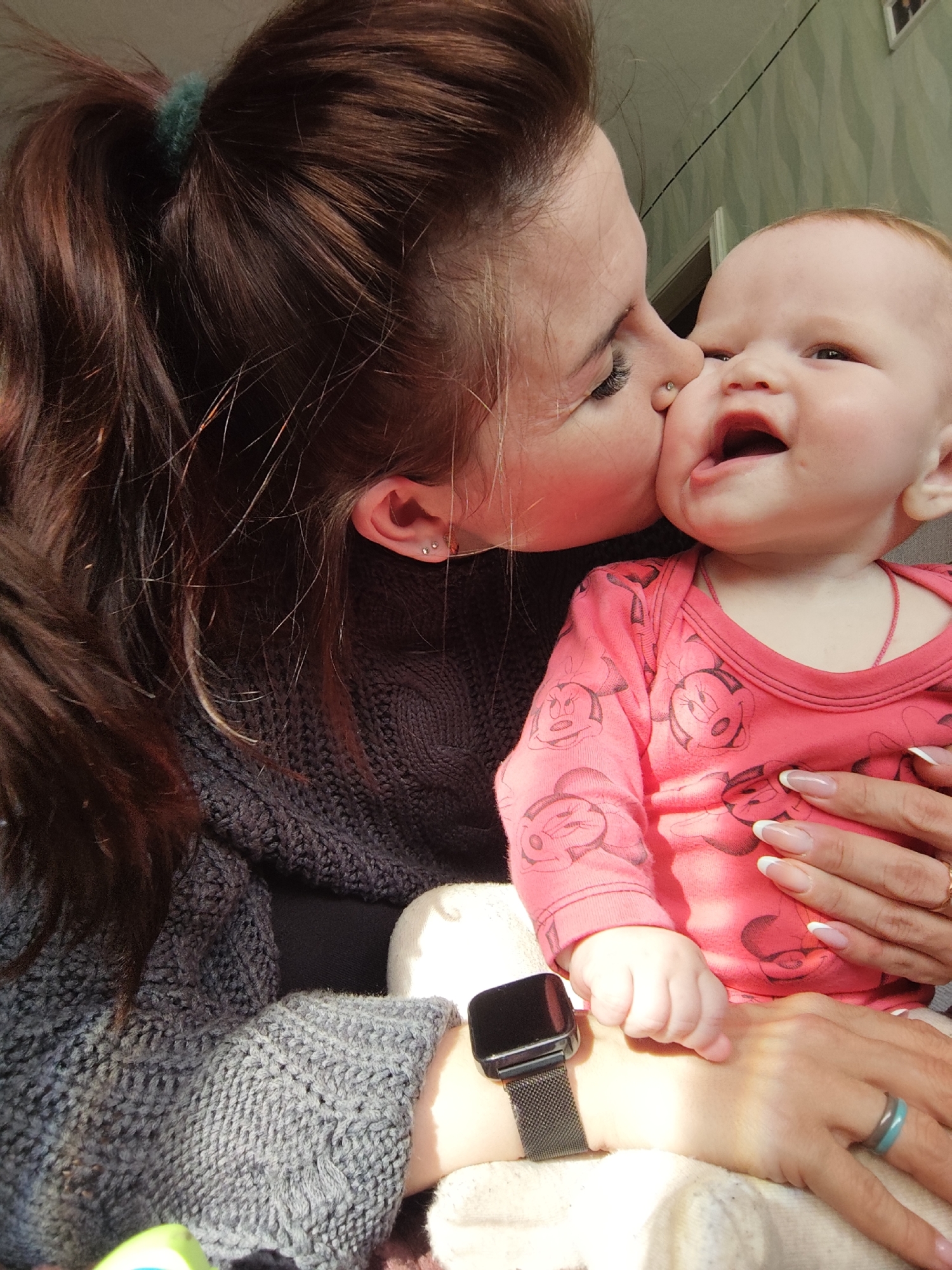 A medium indoor shot shows a woman kissing a baby. The woman has dark brown hair pulled back with a green scrunchie. She is wearing a dark gray knitted sweater, has multiple piercings in her ear, and is wearing a black smartwatch on her left wrist. The baby has light skin, light hair, and is wearing a light red onesie with a Mickey Mouse pattern. The woman is holding the baby and has long, manicured nails with white tips. The background is blurry and shows a wall and doorway. The location is Фоки, Russia.