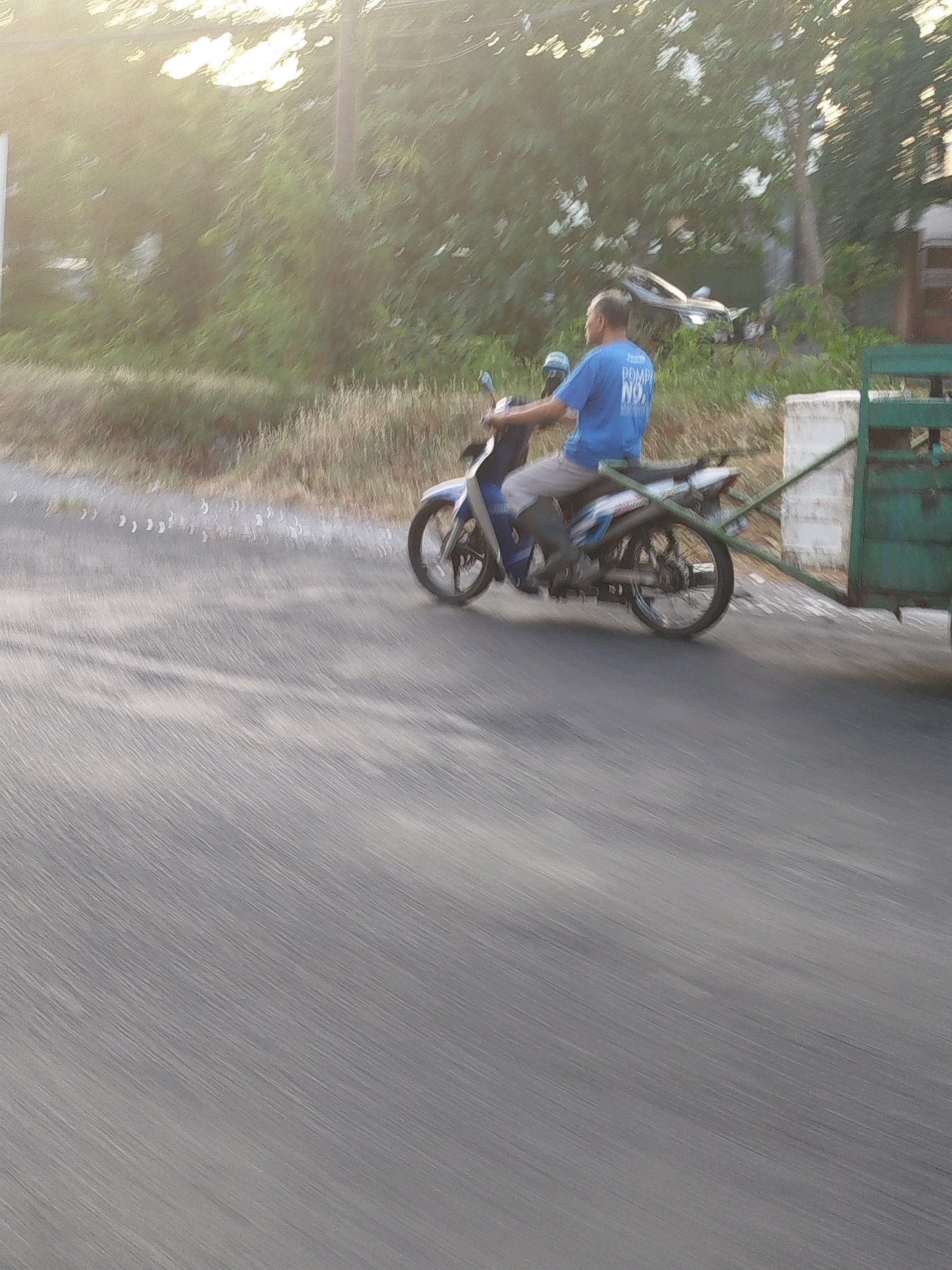 A medium shot captures a man in a blue t-shirt with text on the back riding a motorcycle with a trailer attached. The man, viewed from the side, is wearing light-colored pants and appears to be looking forward. The motorcycle is blue and white. The trailer, green, is partially visible. The scene takes place outdoors on a paved road, with dry grass and trees lining the right side of the road. The sun is setting, casting a warm, hazy light over the image and creating motion blur due to the moving subjects and possibly camera shake, suggesting it is likely sunset. The overall impression is of a rural or suburban setting. The specific location in Surabaya, Indonesia, is not discernable from the image content alone.