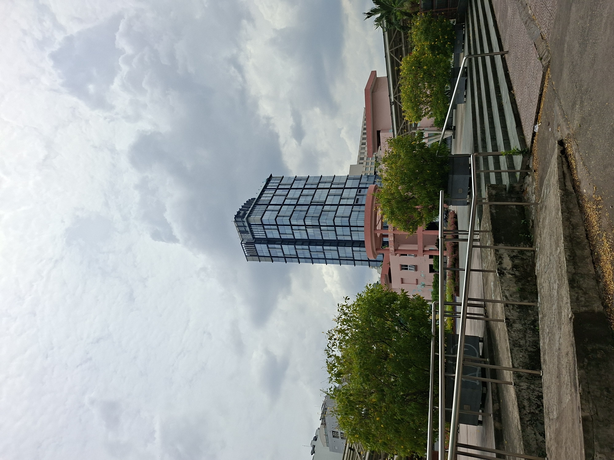 The image depicts an outdoor scene in Thủ Đức City, Vietnam, under an overcast sky filled with a mix of dense and wispy clouds. In the background, a modern, multi-story glass-fronted skyscraper stands tall, its facade reflecting the cloudy sky. In front of the skyscraper, there is a low-rise pink building with visible windows and signage, partially obscured by lush green trees. To the right of the pink building, a portion of another building with a light-colored facade and a patterned roof structure is visible.
In the foreground, a series of concrete stairs with metal railings leads up towards the pink building. These stairs are partially covered with a textured pavement pattern on the right side. Several large, rectangular planters containing green trees with yellow flowers are placed along the top of the stairs. The ground area in front of the stairs is paved with asphalt, with scattered fallen yellow leaves. The overall scene suggests an urban public space, possibly a plaza or an area adjacent to a university campus, given the presence of green spaces and modern architecture. No people are visible in the frame.