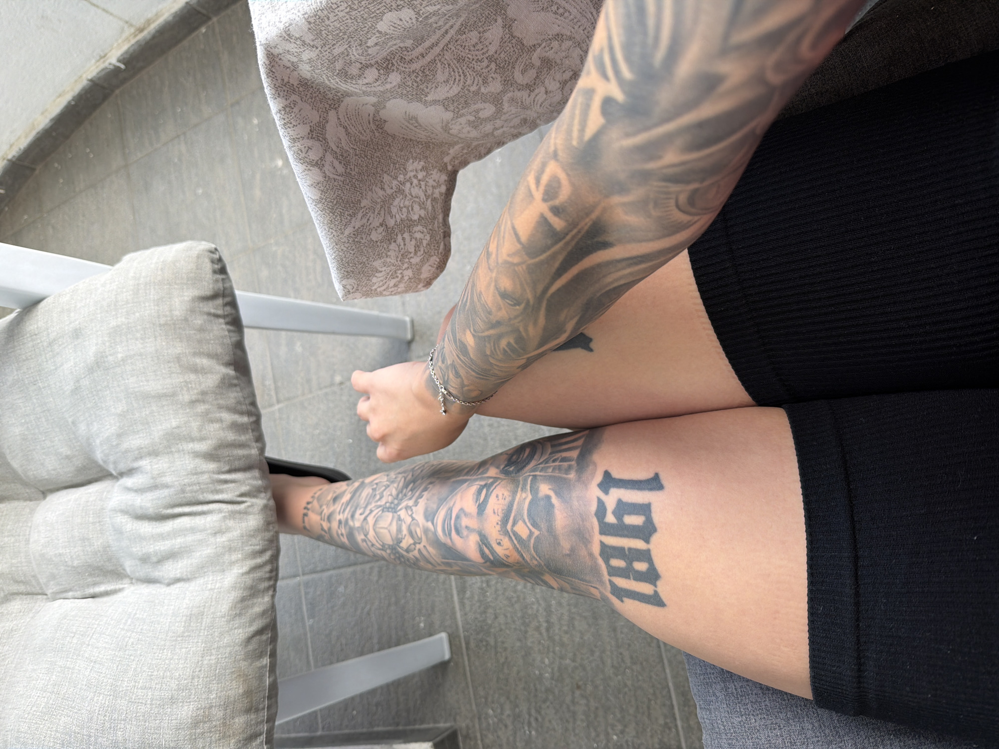 A person's legs and arm are visible, adorned with extensive black ink tattoos. The tattoos on the left leg depict a large, detailed face with elaborate headdress, and the number "1861" in a gothic font. The right arm also has a full sleeve of intricate tattoos. The person is seated on a grey cushioned chair, with a grey, patterned tablecloth visible on a table to the right. The background consists of a tiled floor, suggesting an outdoor setting. The lighting suggests daytime. There are no visible identifying landmarks or specific urban features that would indicate a location in Plovdiv, Bulgaria.