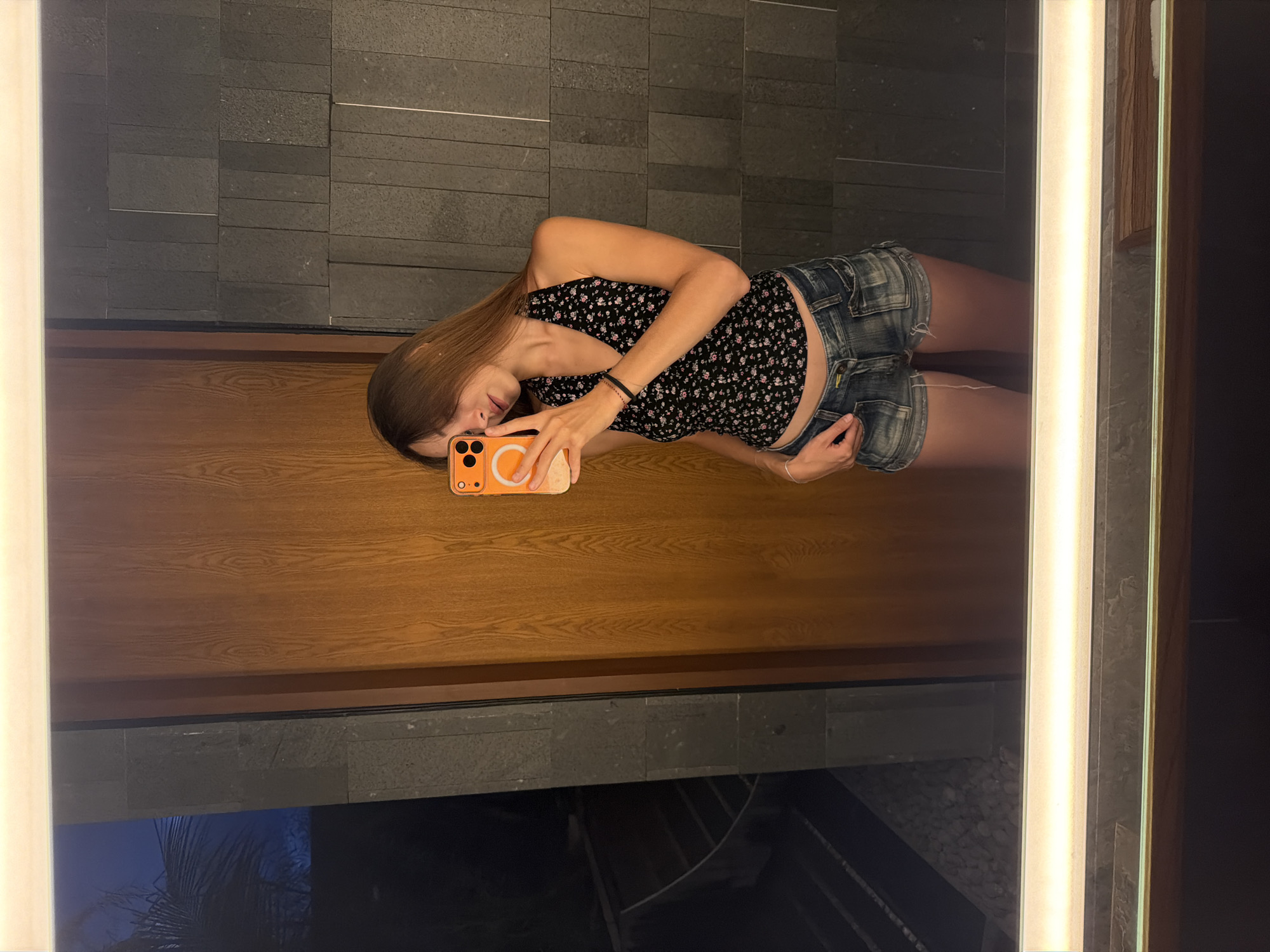 A young woman stands in front of a mirror and takes a selfie. She has long, light brown hair. She is wearing a sleeveless black top with small floral patterns and denim shorts. She holds an orange smartphone in her left hand and is posing in front of the mirror. The mirror reflects a modern interior with a dark gray stone wall, a brown wooden panel, and an illuminated light strip on the mirror frame. The lower part of the mirror reflects a night-time scene with foliage, suggesting the location might be near a garden or outdoor area. The architectural style and setting suggest the photo was taken in the Ubud District of Indonesia, where modern design is often integrated with natural elements.